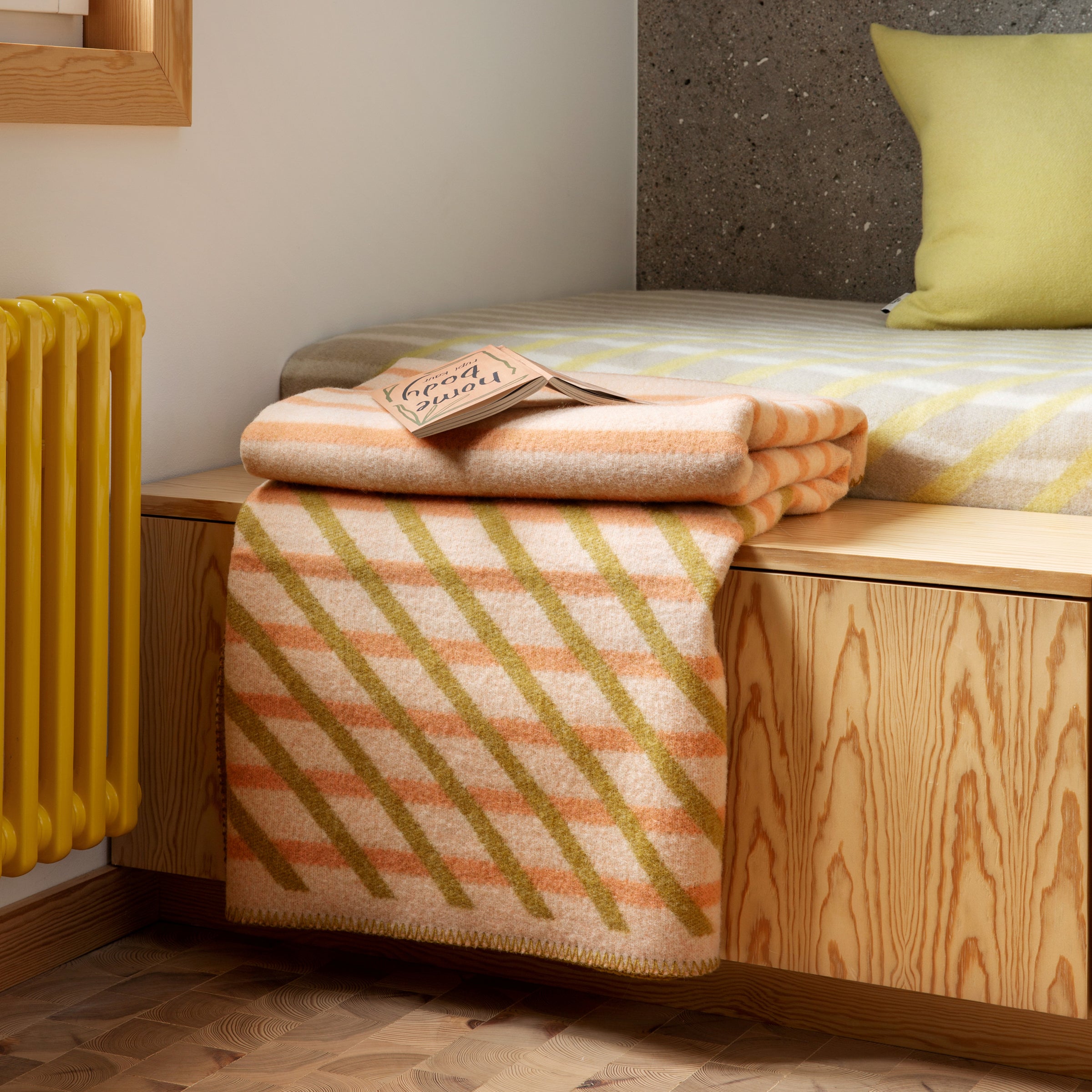 A cozy bedroom scene features the BISLETT Throw by Roros, a folded, diagonal-striped blanket made from Norwegian lambswool in peach and green draped over a wooden bench. A matching pillow and throw adorn the bed, complemented by a yellow radiator and wooden herringbone floor for added warmth.
