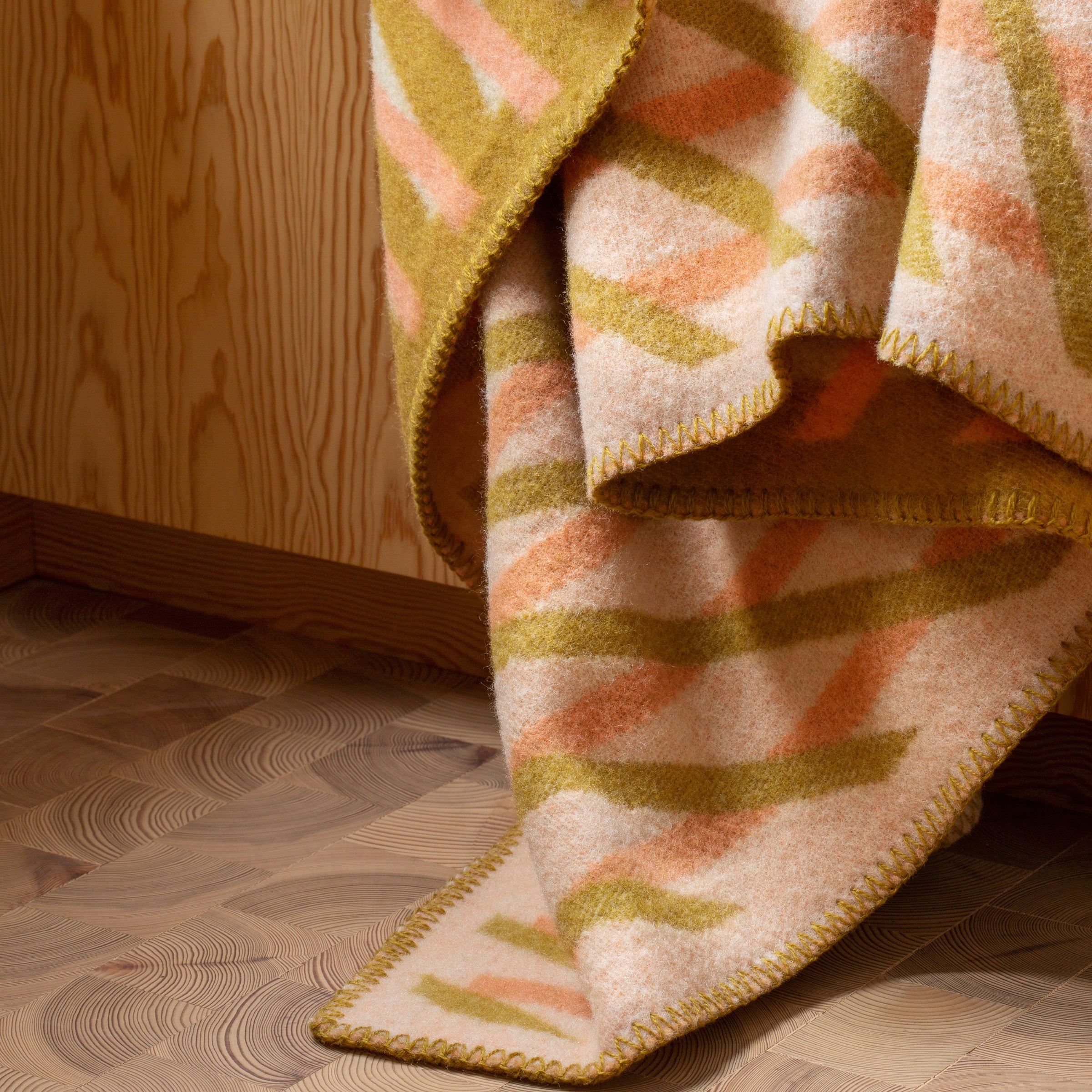 The BISLETT Throw by Roros, made of folded Norwegian lambswool with an abstract green, orange, and beige pattern, elegantly drapes over a wooden surface. The wood floor and wall enhance its cozy warmth.