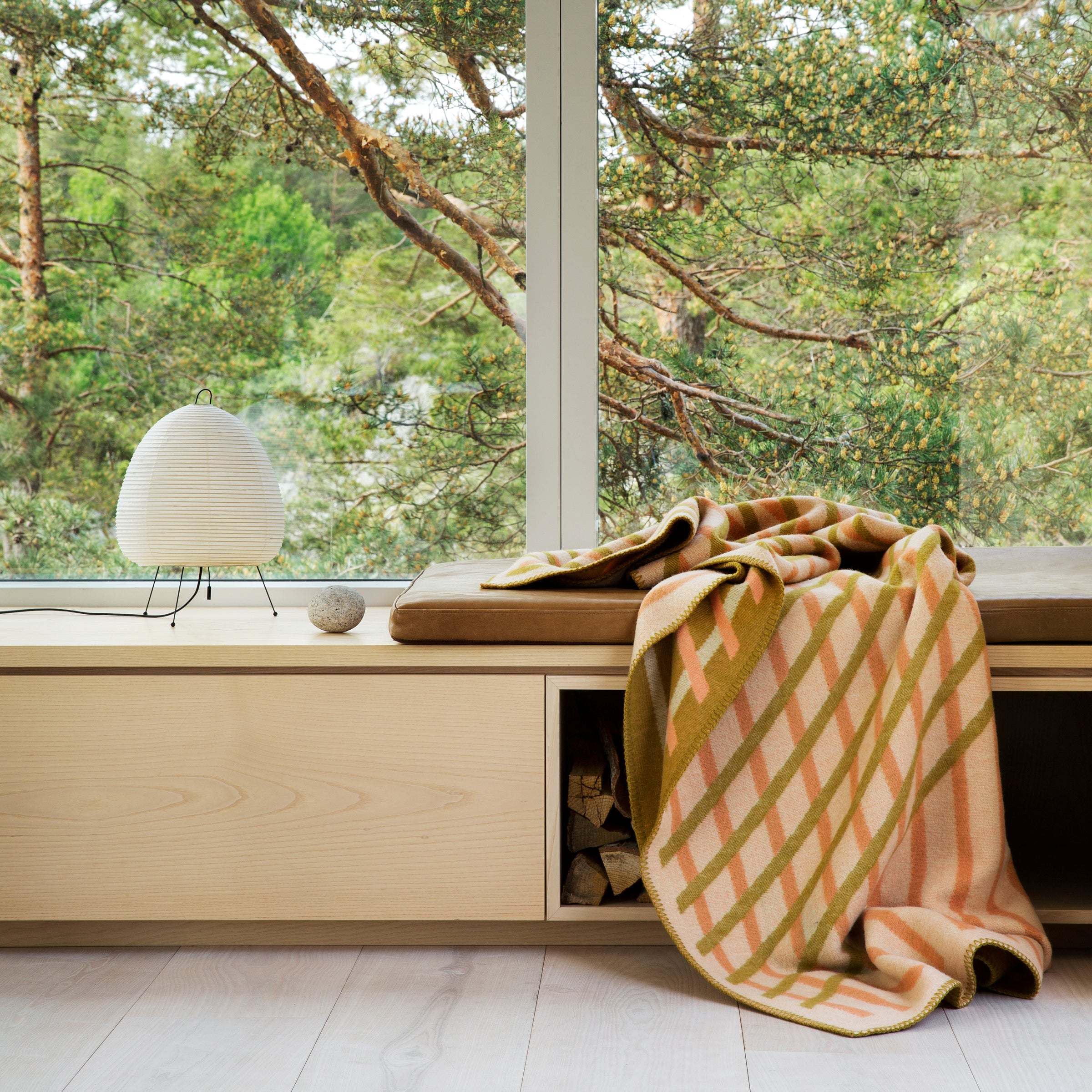 A scenic forest view is seen through a large window. Inside, a wooden bench with a cushion features the orange and green striped BISLETT Throw by Roros. A small white lamp and a stone rest on top, while logs are neatly stored in the shelf beneath.