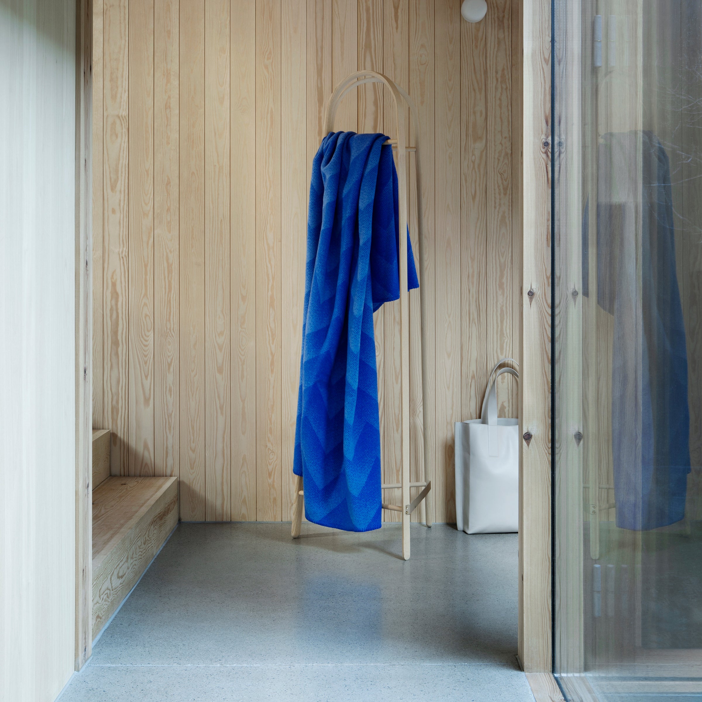 A Lynild Throw by Roros, echoing Scandinavian charm, hangs on a wooden stand in a room with light wood-paneled walls and a concrete floor. A white bag rests beside it, enhancing the minimalist spaces serene vibe.
