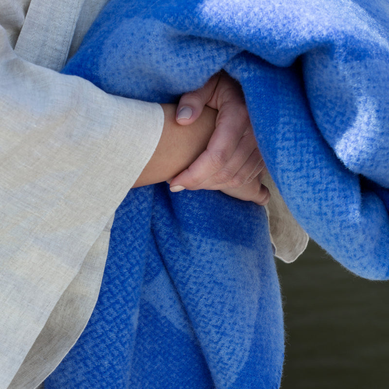 A person in a light garment gently clasps their hands under the Lynild Throw, a thick blue blanket made of Norwegian lambswool by Roros, softly lit by sunlight reminiscent of Anderssen & Volls elegant craftsmanship.