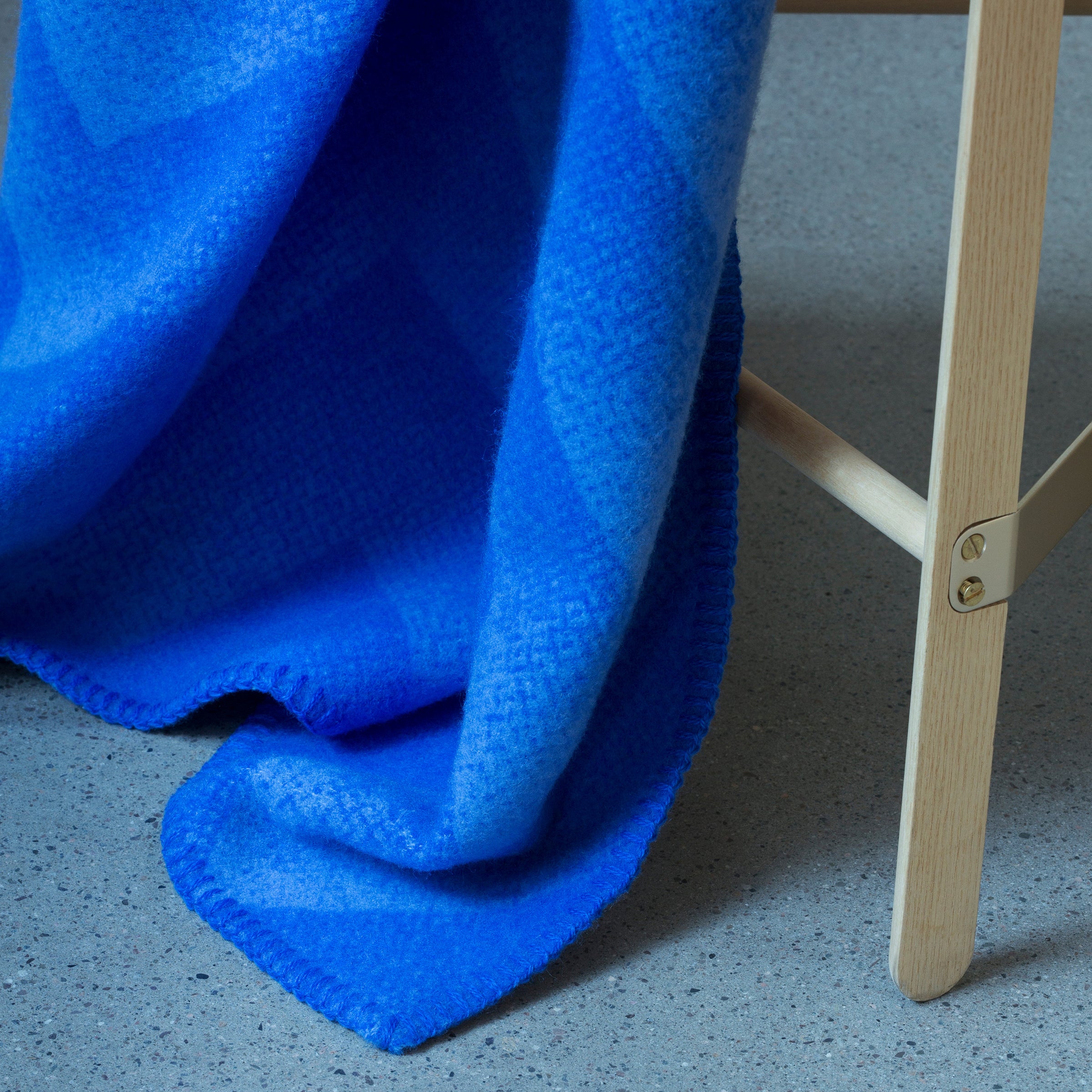 The Lynild Throw by Roros, in vibrant blue and made from luxurious Norwegian lambswool with a subtle geometric pattern, elegantly draped over a wooden chair on a gray textured floor.