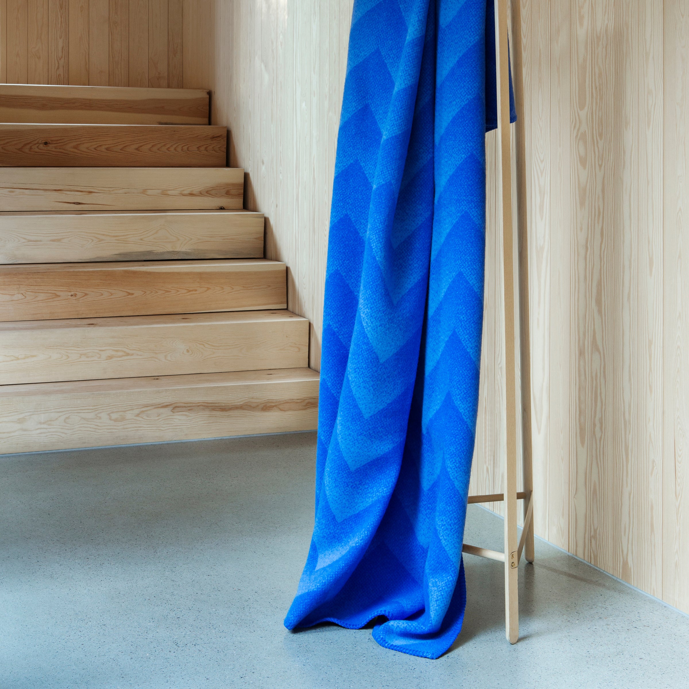 The Lynild Throw by Roros, a vivid blue blanket crafted from Norwegian lambswool with a fire stitch pattern, elegantly drapes over a wooden stand in a minimalist room with light wood walls and staircase, and polished concrete floors.