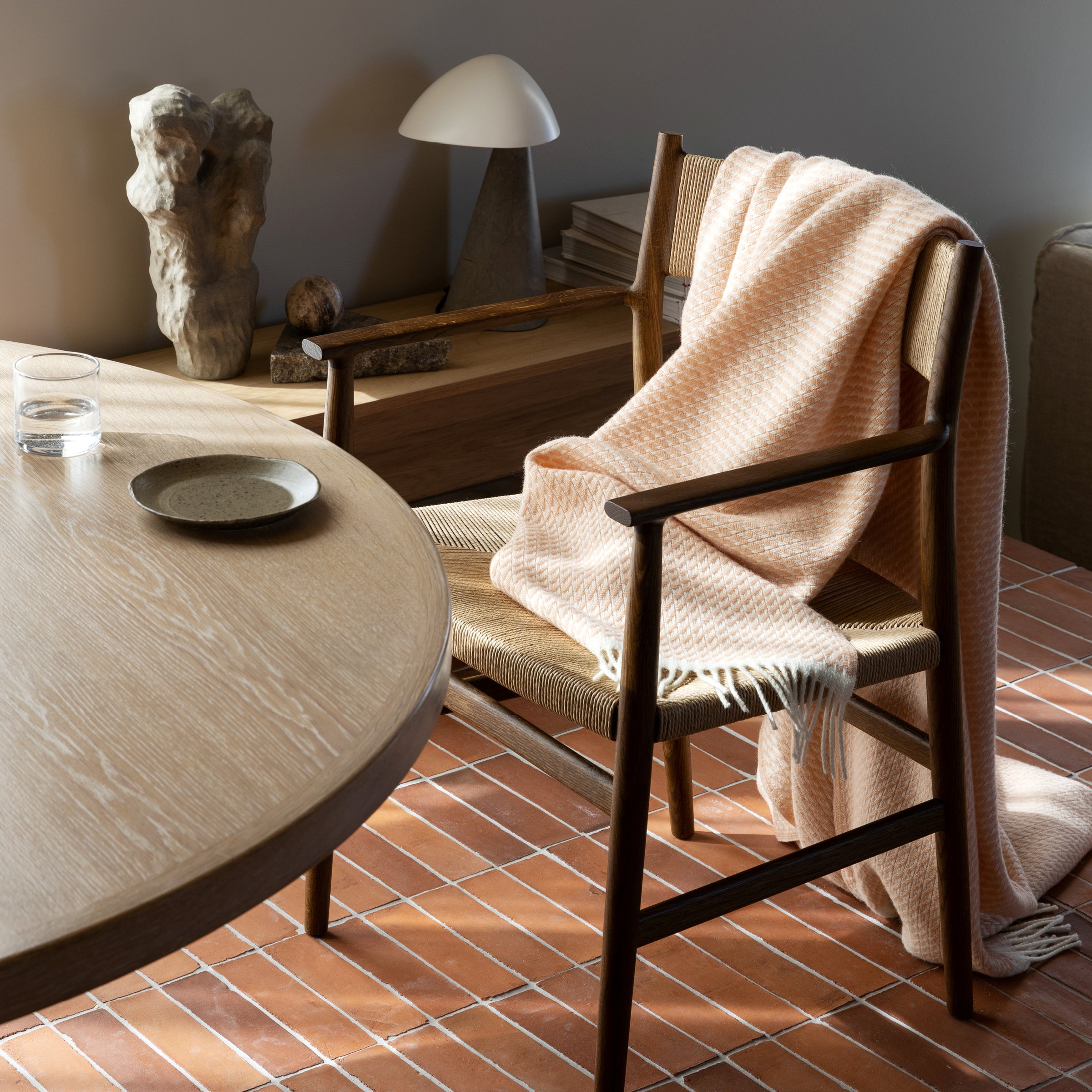 A cozy room features a wooden chair draped with a MELLO Throw from Roros, next to a round wooden table with a glass and plate. A sculptural lamp and decor pieces enhance the woolen interiors on the tiled floor.