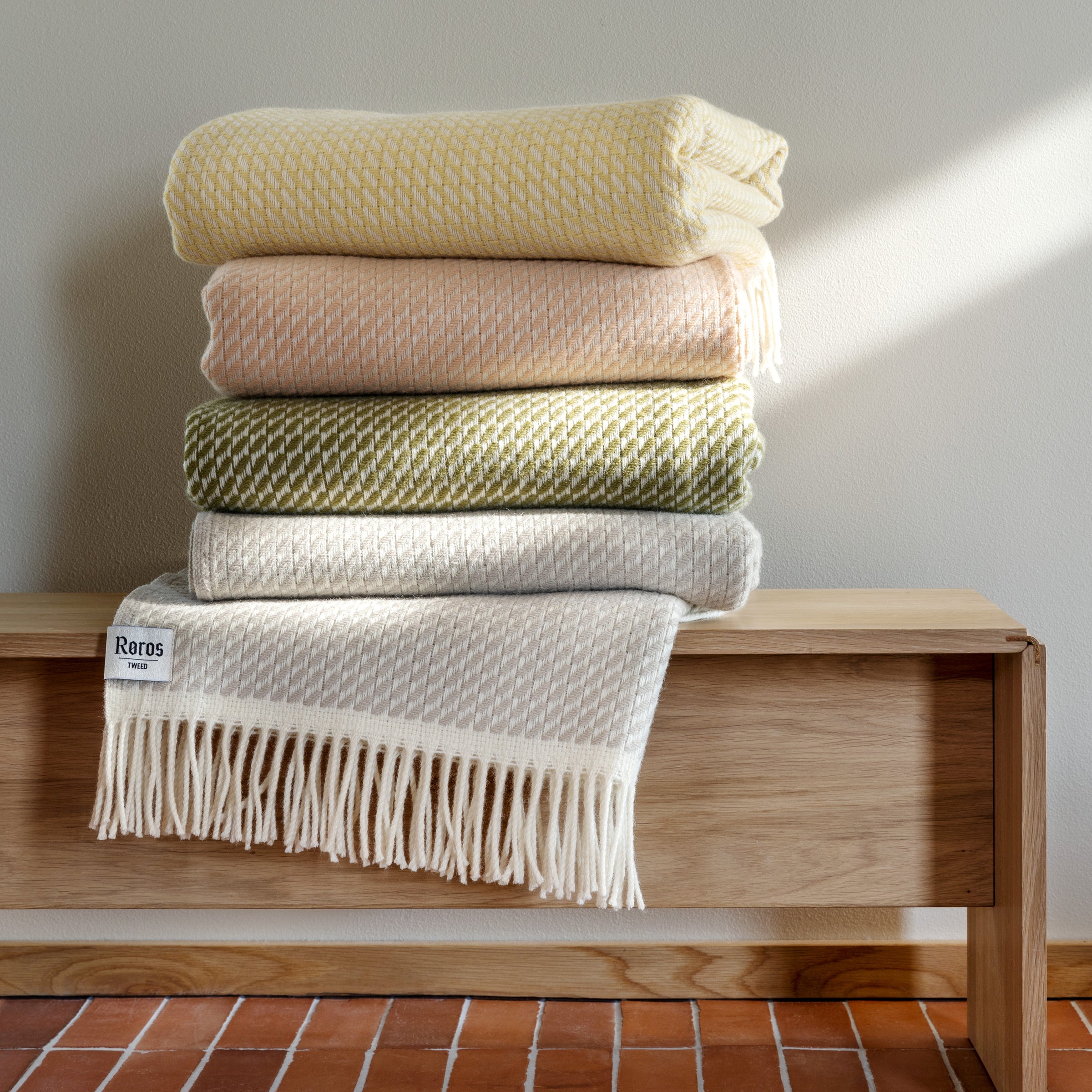 A stack of Roros MELLO Throws in yellow, peach, green, and beige rests on a wooden bench against a light gray wall. The bottom throw, made from Norwegian lambswool, features a fringe and tag. Sunlight casts cozy shadows, creating inviting woolen interiors.