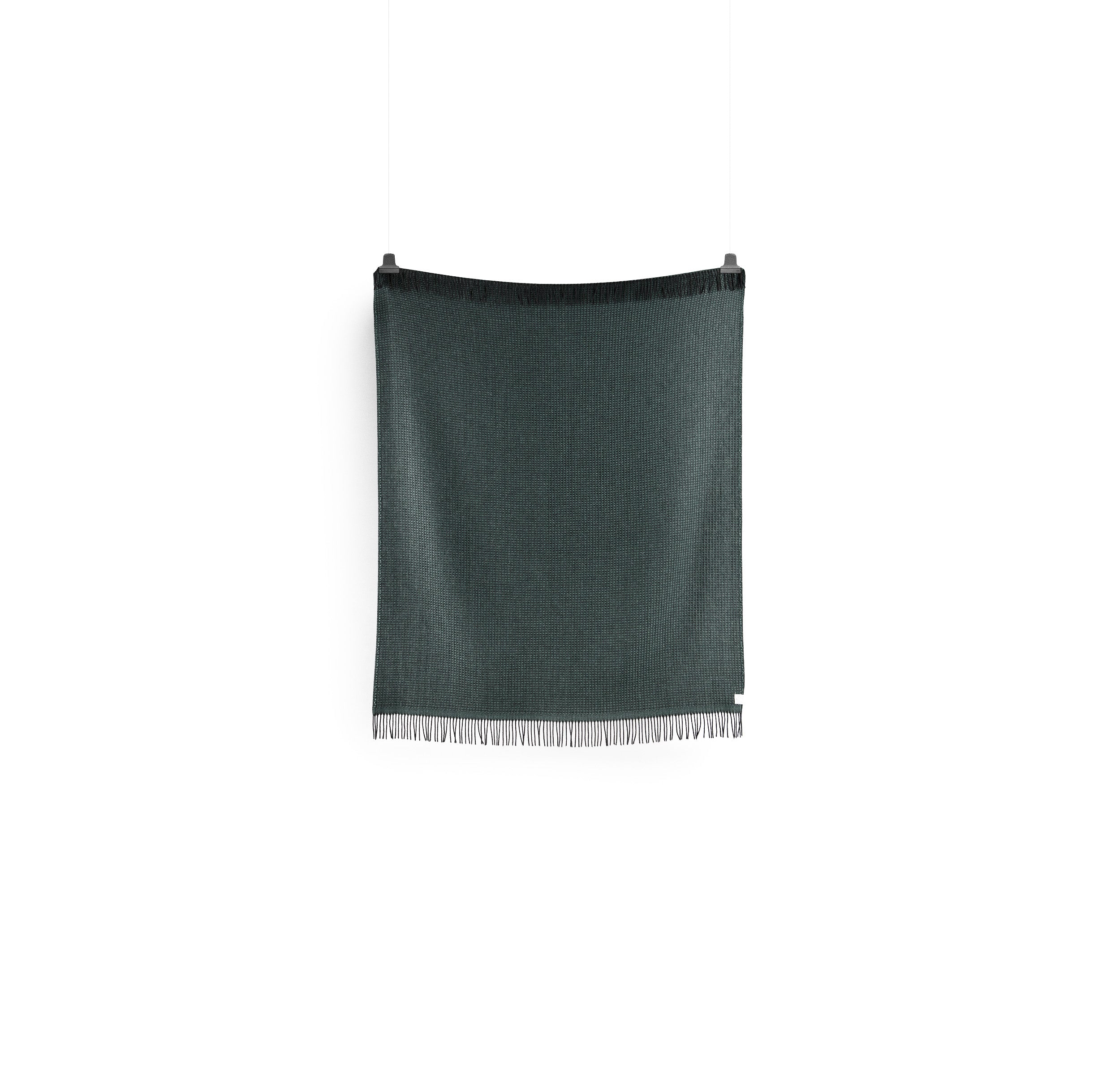 The VEGA throw by Roros, in a dark green reminiscent of Norwegian lambswool, hangs against a white background with a subtle texture. Its suspended by two visible clips at the top, and the bottom edge is accented with fringes or tassels.