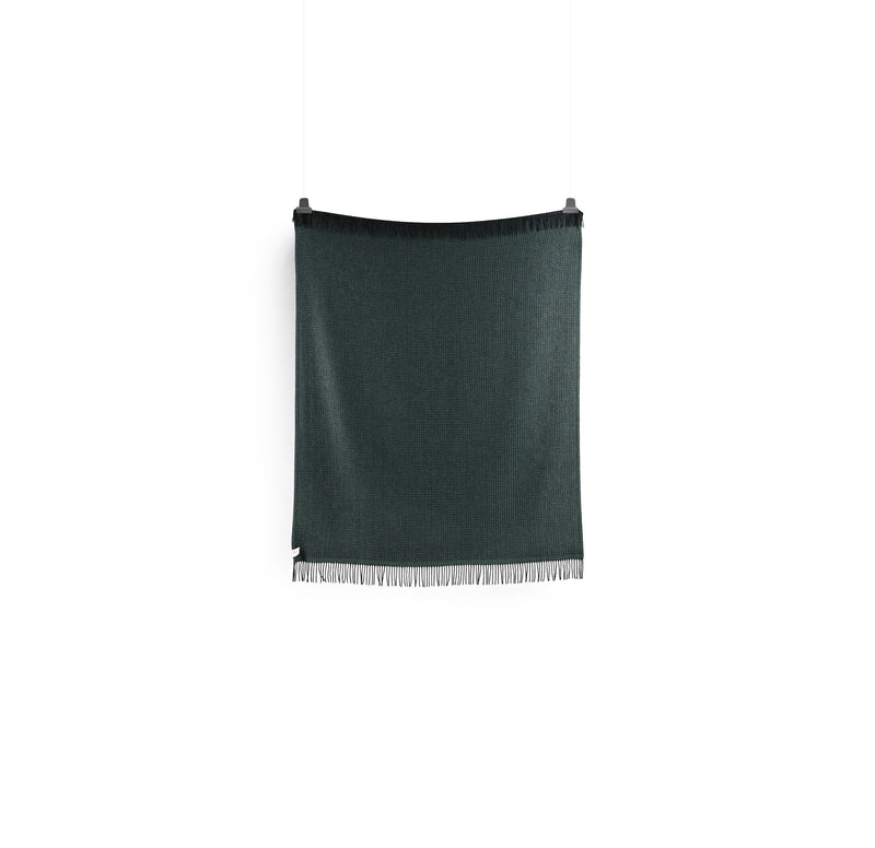 The VEGA Throw by Roros, designed by Anderssen & Voll, elegantly hangs against a white background. Its crafted from Norwegian lambswool and features a dark green woven textile with fringe.