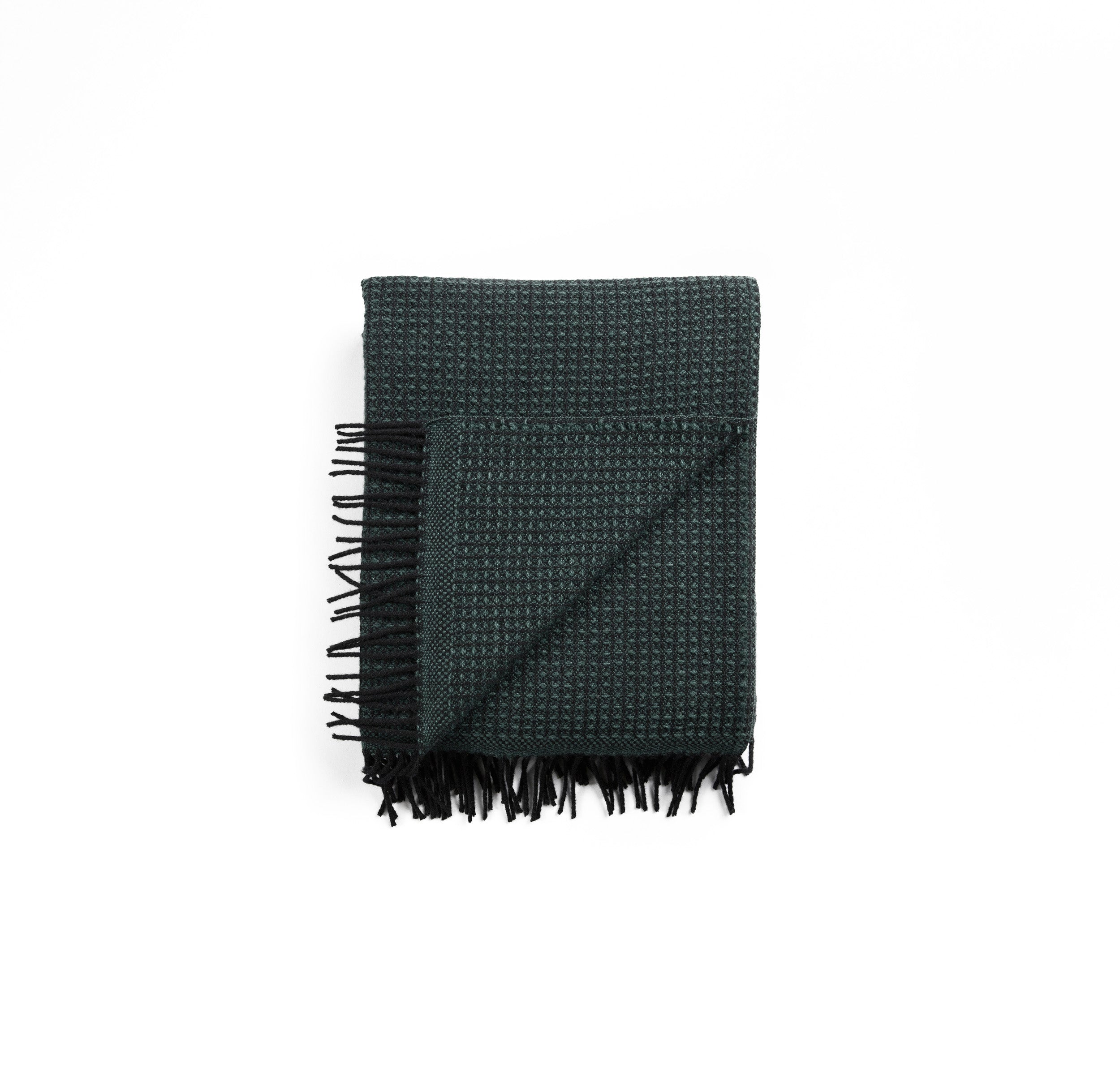 The VEGA Throw by Roros is crafted from Norwegian lambswool and features a folded dark green textured design with fringe on the edge against a white background.