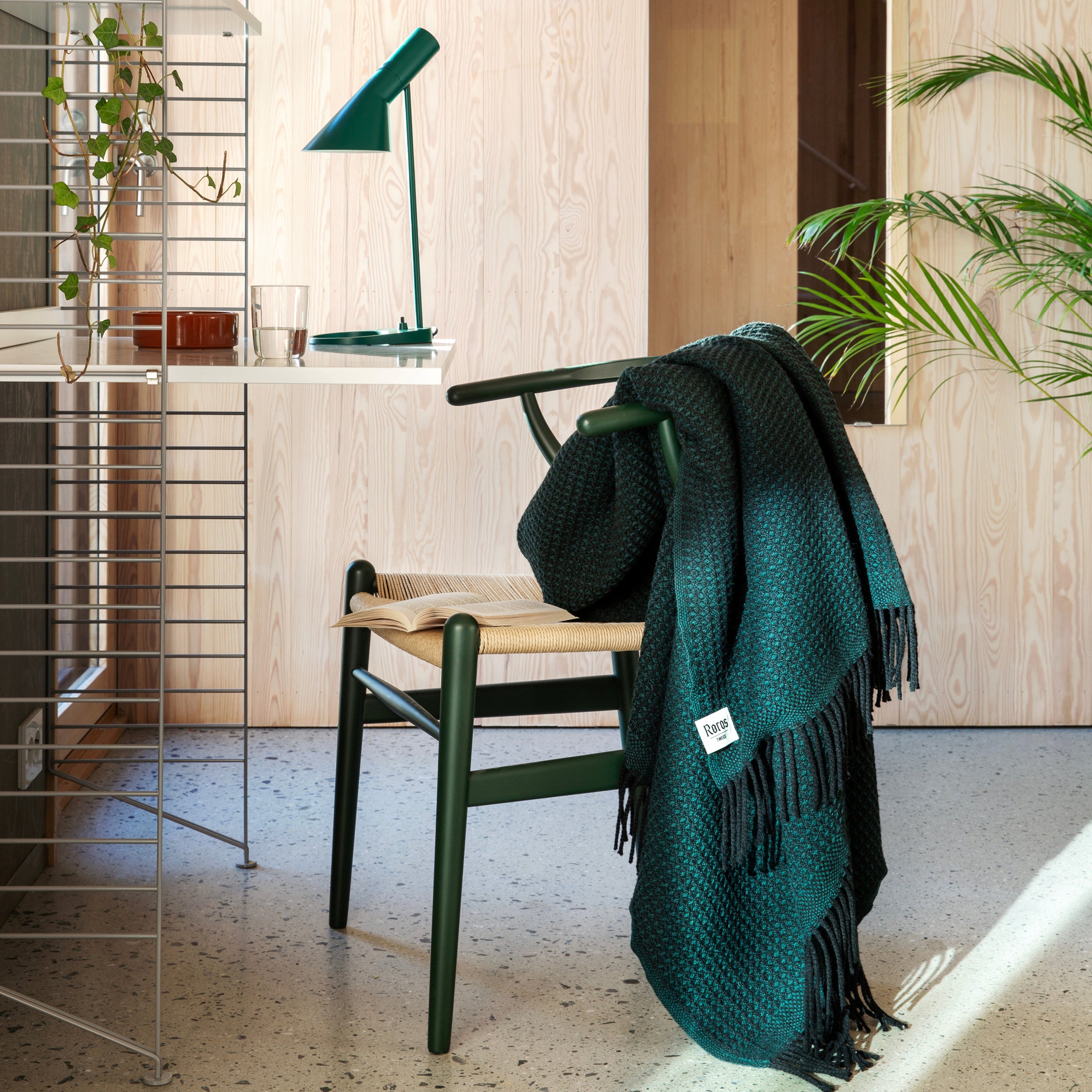 A cozy room features a green chair with a Roros VEGA throw crafted from Norwegian lambswool. A green desk lamp and potted plant accompany it, while light wood-paneled walls and a large leafy plant in the background complete the inviting setting designed by Anderssen & Voll.
