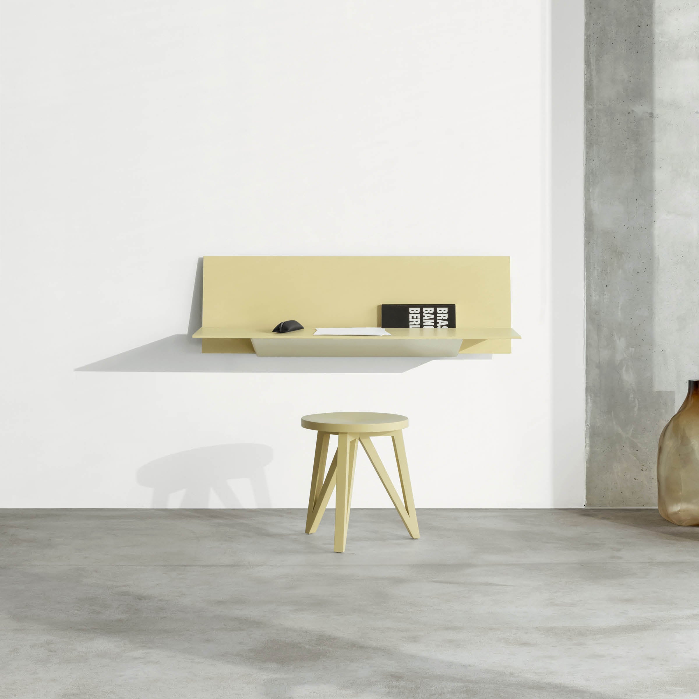 A minimalist workspace showcases the Loehr S1 CHAPTER Bureau, a light yellow wall-mounted floating desk paired with a matching stool. The desk holds a closed laptop and a small book, while a large ceramic vase sits on the concrete floor, highlighting sleek, stylish wall-mounted furniture.