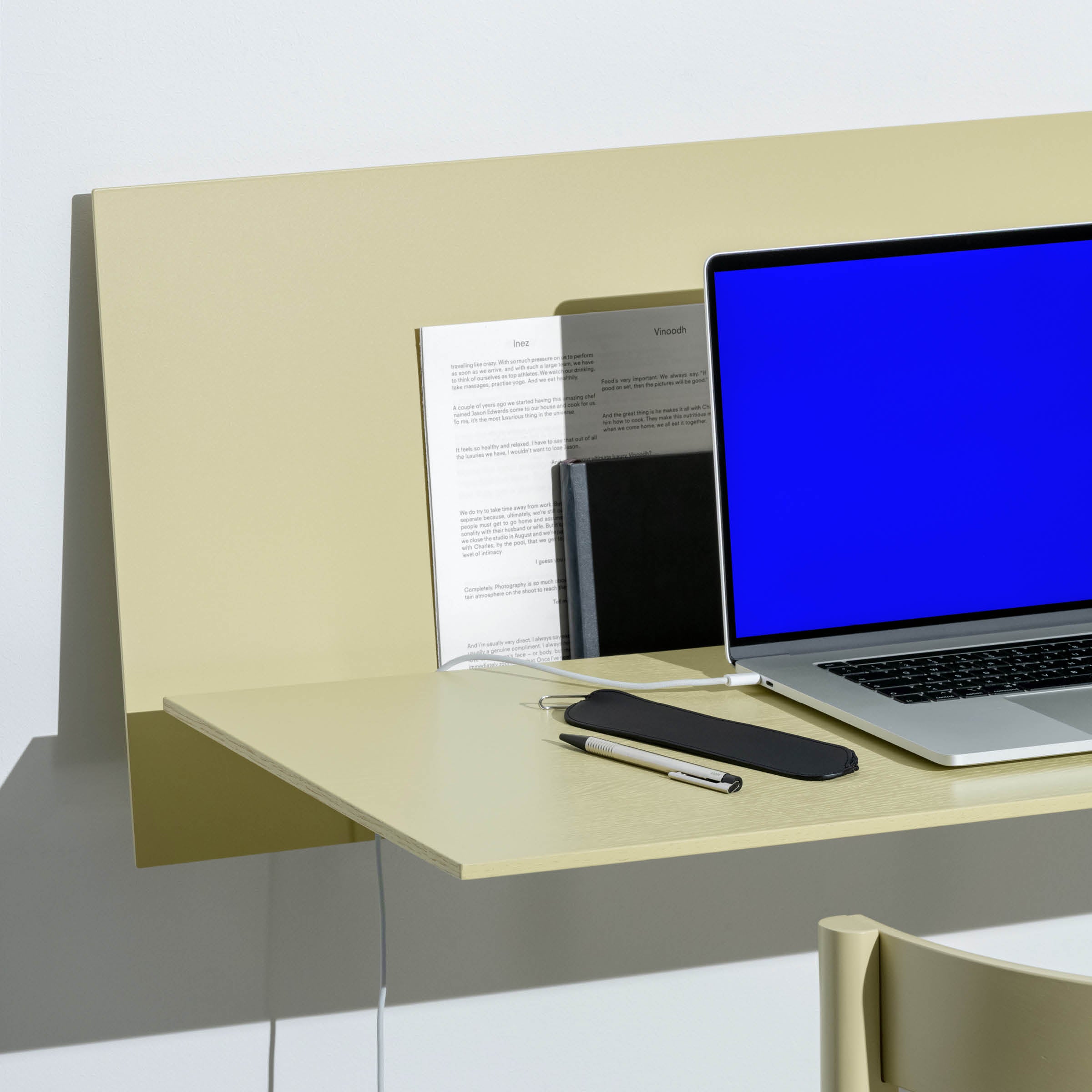 A minimalist desk setup showcases the S1 CHAPTER Bureau by Loehr, a beige standing desk with a sleek laptop showing a blue screen. Accessories include a pen, smartphone, organized papers, and part of a light wooden chair. Bright lighting highlights the clean modern aesthetic.