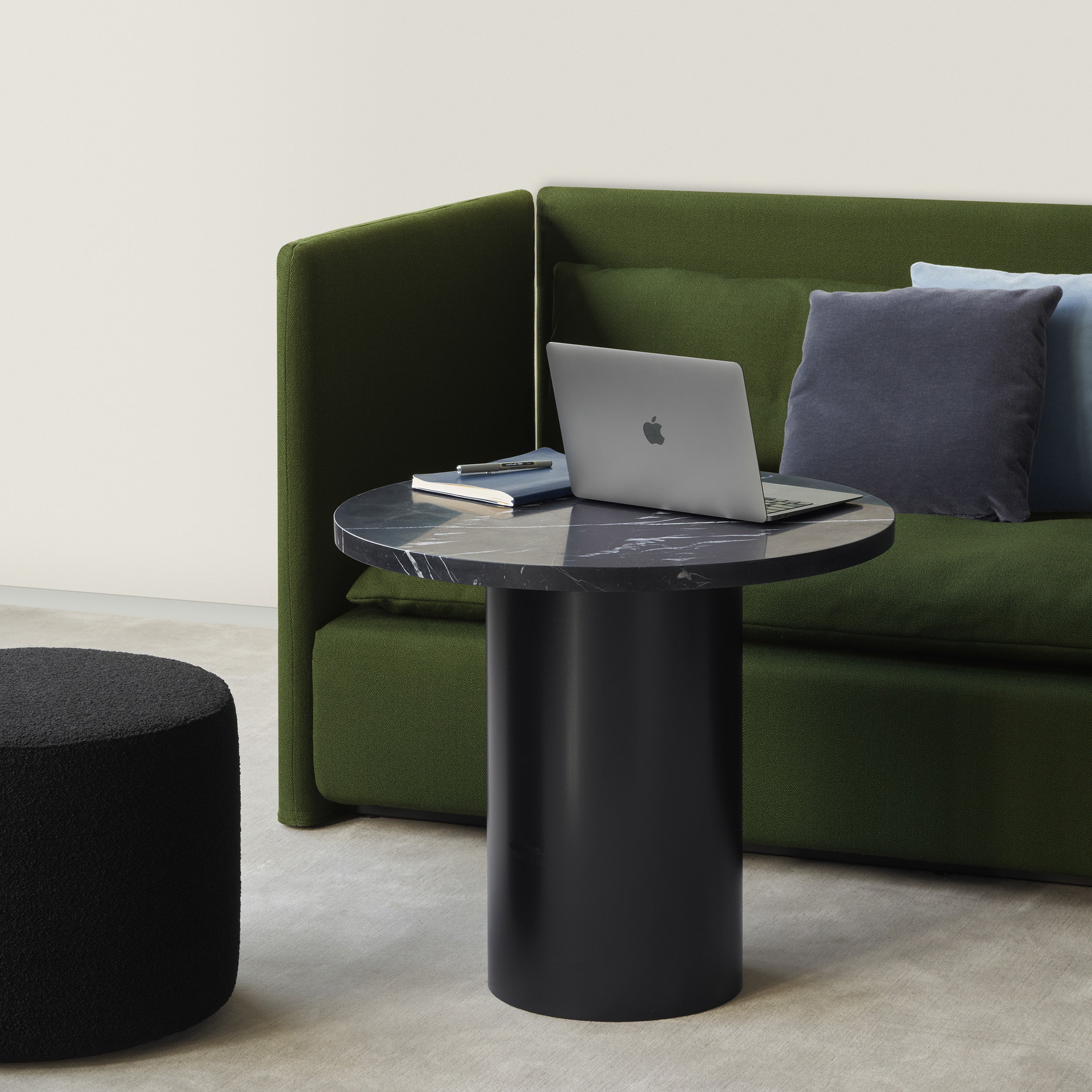 A modern room includes a green sofa with blue and gray cushions. A sleek E15 CT09 ENOKI table, showcasing rich marble, supports a closed laptop and books. In front of the sofa, a black ottoman sits on the light-colored carpet.