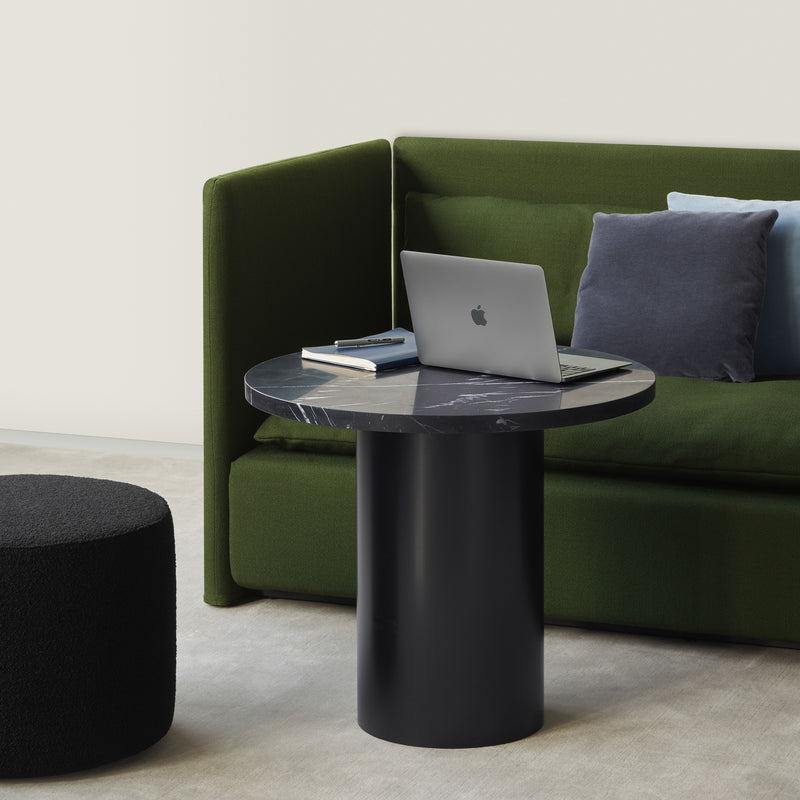 A modern room includes a green sofa with blue and gray cushions. A sleek E15 CT09 ENOKI table, showcasing rich marble, supports a closed laptop and books. In front of the sofa, a black ottoman sits on the light-colored carpet.