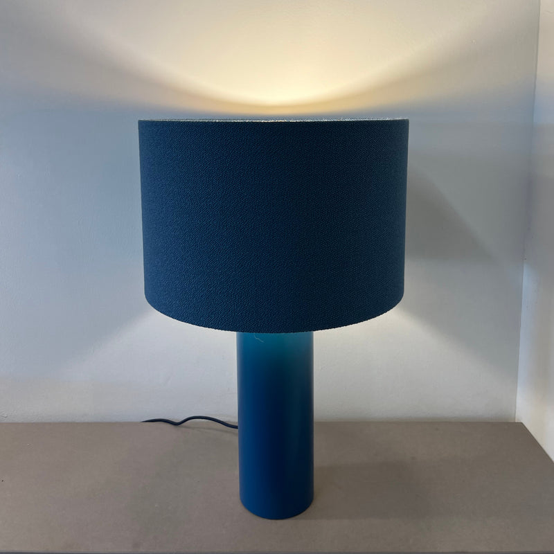 The ALL ROUND Table Lamp by Victor Foxtrot features a blue cylindrical powder-coated steel base and matching drum shade. It sits elegantly on a neutral surface, casting a soft glow on the wall. Currently on sale!.