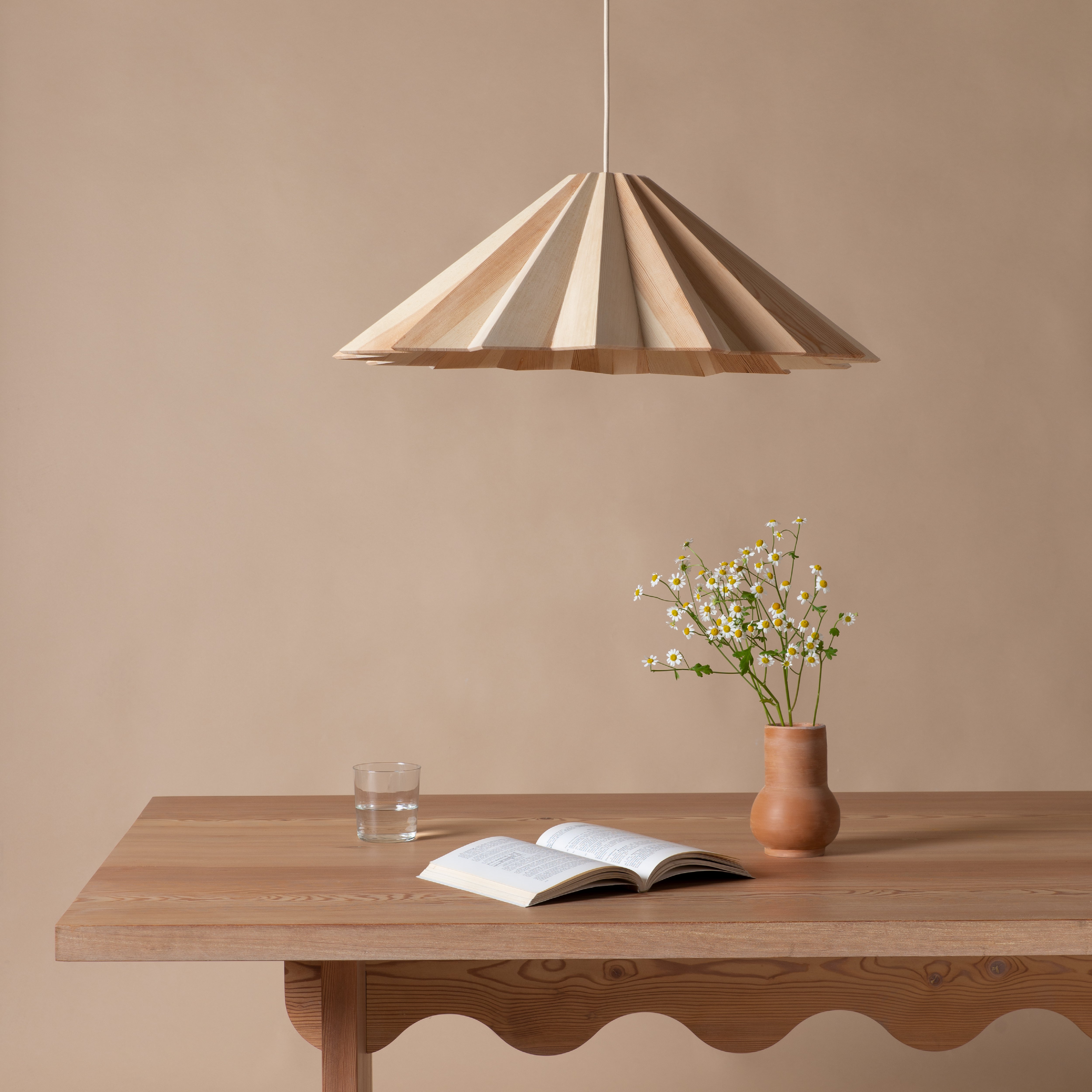 A minimalist scene features a wooden table with an open book, a glass of water, and a clay vase with small flowers. Above, the STRATUS Big lamp by Schneid, made from handcrafted veneer, adds warmth against the neutral beige wall backdrop.