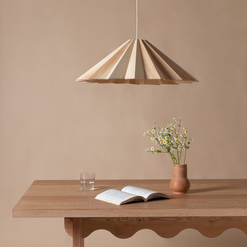 A minimalist scene features a wooden table with an open book, a glass of water, and a clay vase with small flowers. Above, the STRATUS Big lamp by Schneid, made from handcrafted veneer, adds warmth against the neutral beige wall backdrop.