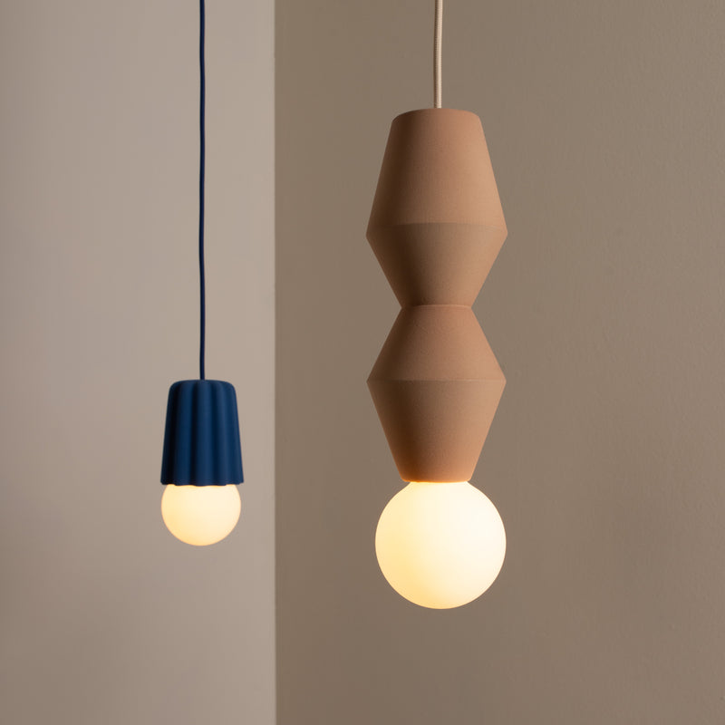 Two pendant lights hang like sculptures: the left features a blue angular shade with a round bulb, exuding creativity; the right, named Stucco No. I by Schneid, displays a tan stacked geometric design with a glowing bulb, set against a neutral beige background.
