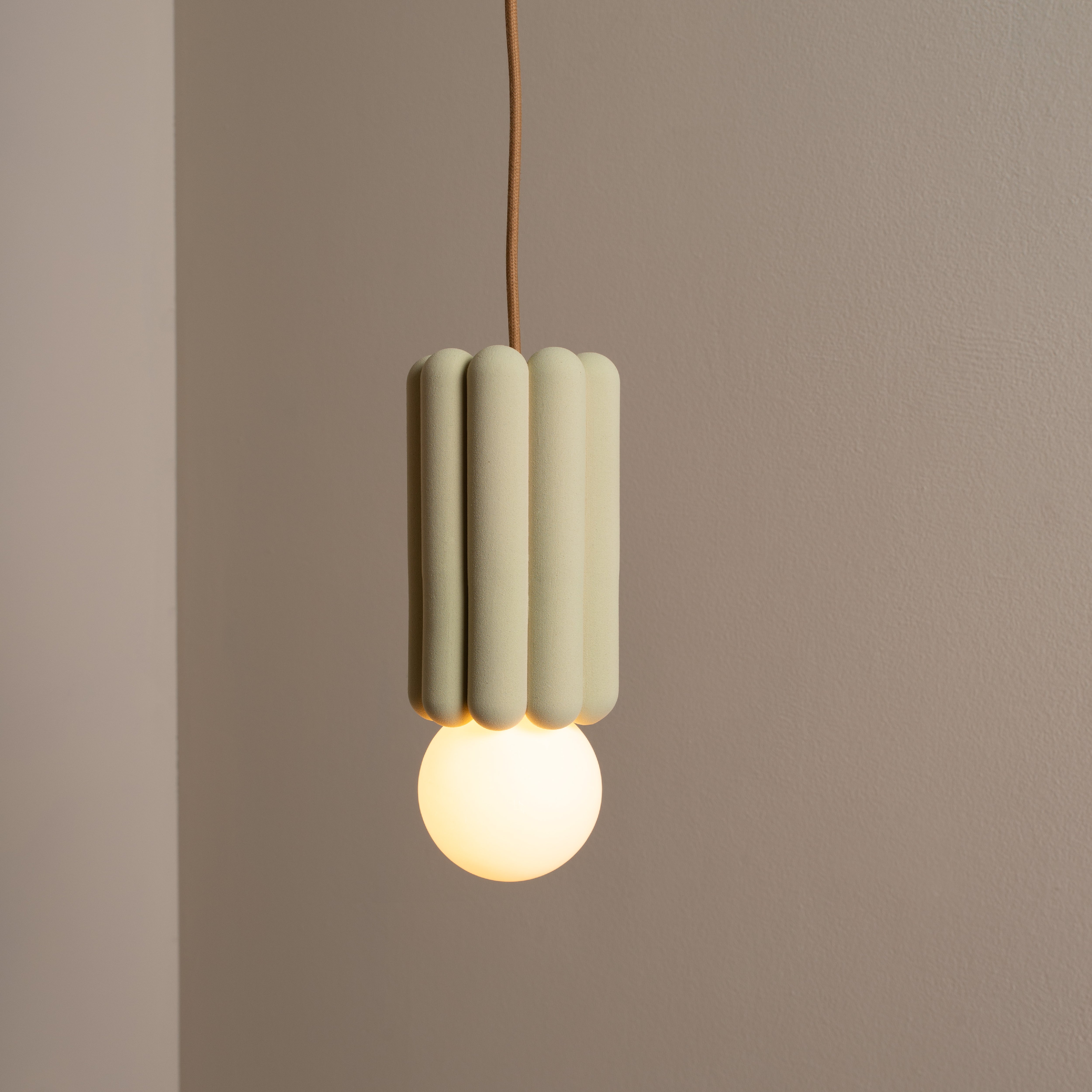 The Stucco No. I by Schneid suspends like a sculpture, featuring a ribbed, cylindrical green shade and glowing round bulb at the bottom, casting creative luminosity on a plain beige wall.