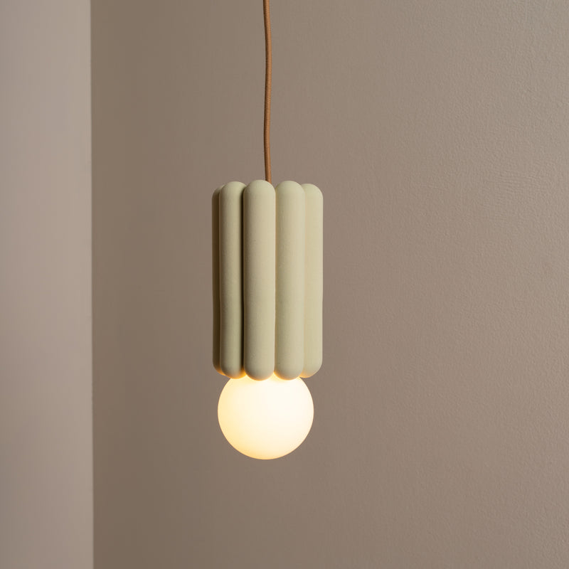 The Stucco No. I by Schneid suspends like a sculpture, featuring a ribbed, cylindrical green shade and glowing round bulb at the bottom, casting creative luminosity on a plain beige wall.