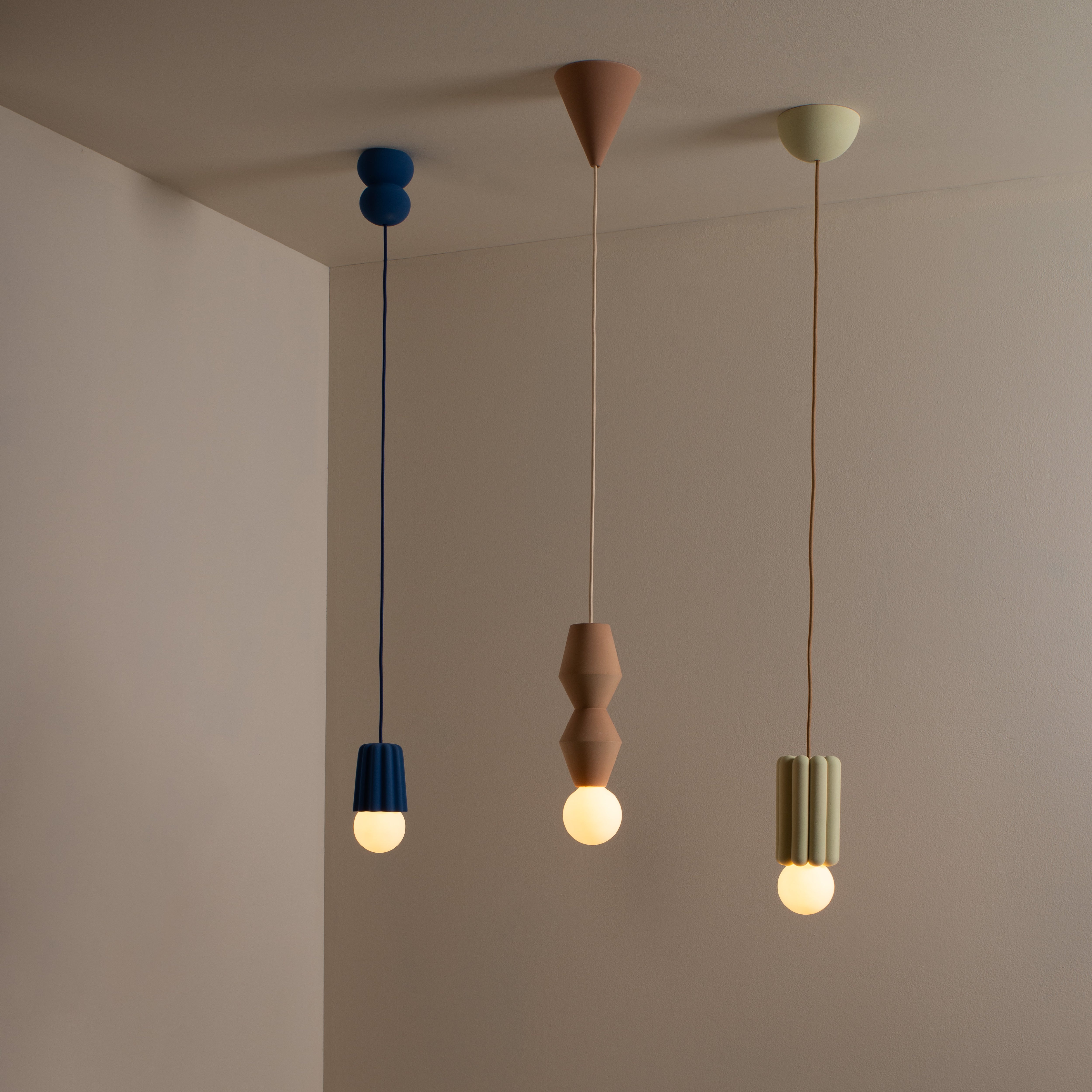 Three Schneid Stucco No. II pendant lights with round bulbs hang from the ceiling, each having a distinct geometric shape and in blue, terracotta, and green. They stand out against the neutral stucco wall in the background. Crafted with ceramic precision, they add charm to any space.
