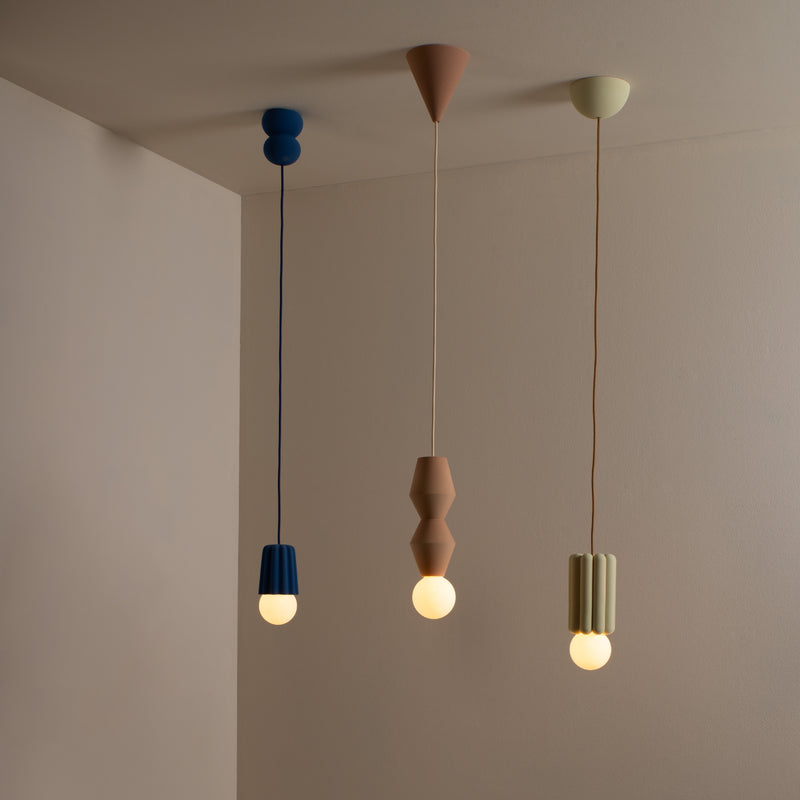 In a minimalist setting, three pendant lights from Schneids Stucco No. I line hang from the ceiling like sculptural pieces, each featuring distinct shapes and colors: round blue elements, geometric brown shapes, and pale green vertical ridges; together they cast a creative luminosity.
