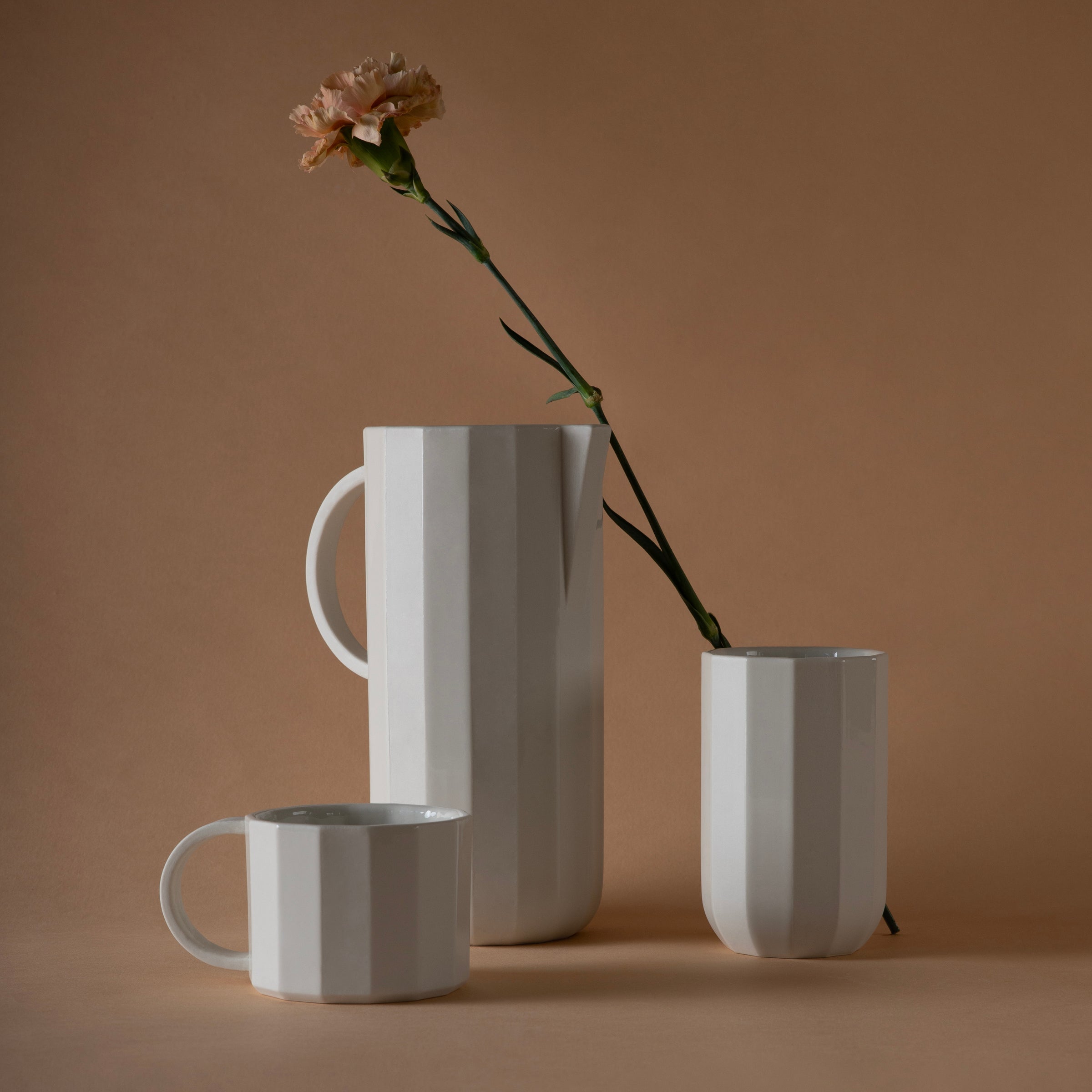 A still life features Schneids Facet Tableware, showcasing a white mug, jug, and tall cup with vertical ridges against a brown backdrop. A pink carnation in the jug adds elegance and highlights the subtle color dynamics.