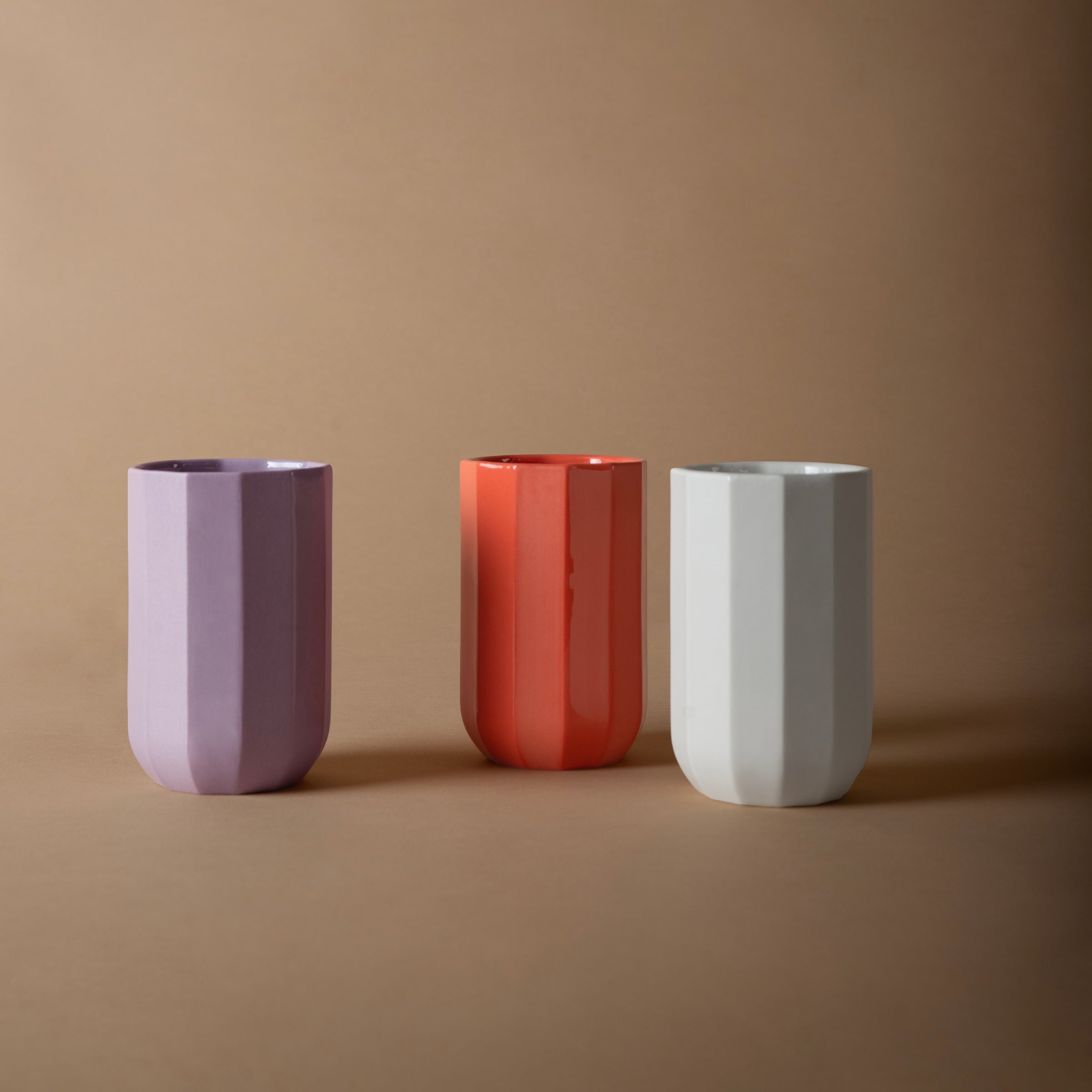Three geometric Facet Tableware cups by Schneid are placed on a brown surface against a matching background. The ceramic cups are arranged from left to right in purple, orange, and white, showcasing their unique charm.