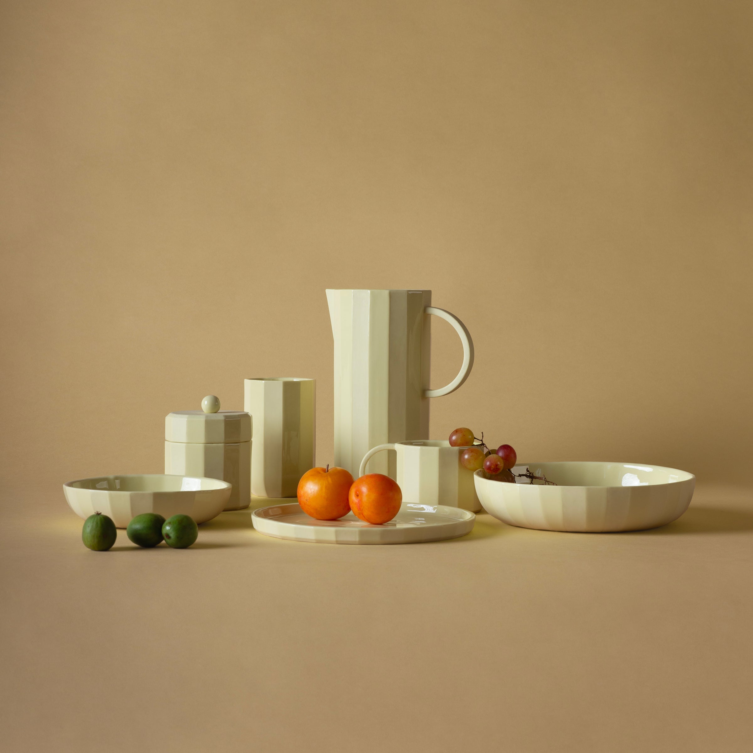 The Facet Tableware collection by Schneid, featuring cream-colored ceramic elements like a small bowl, is elegantly arranged on a beige background. Vibrant fruits such as oranges, grapes, and green plums add color and texture to the sophisticated display.