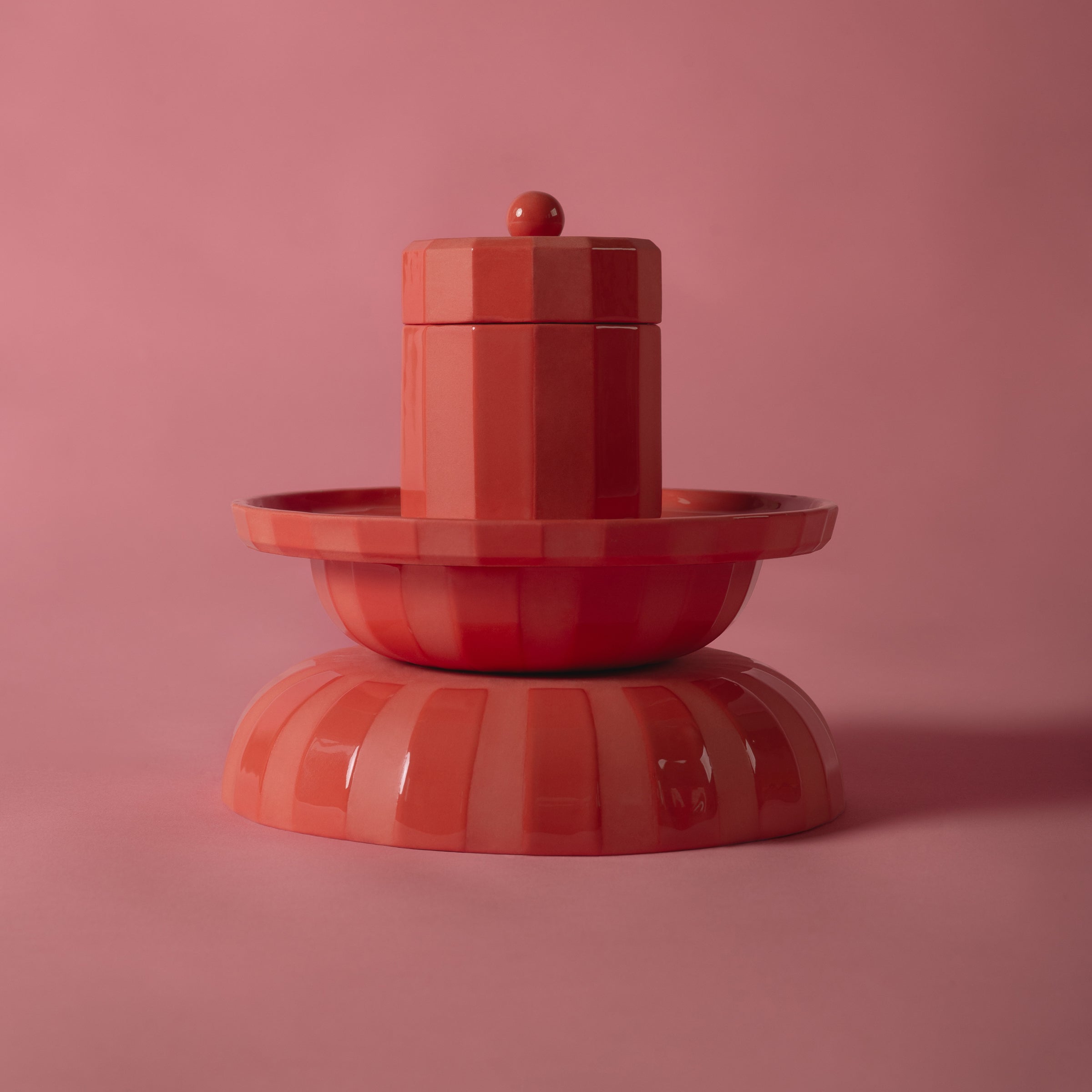 A glossy red ceramic item with geometric shapes, resembling a decorative fountain, rests on a matching surface. The soft red background enhances the shine and elegance of Schneids Facet Tableware - Plate.