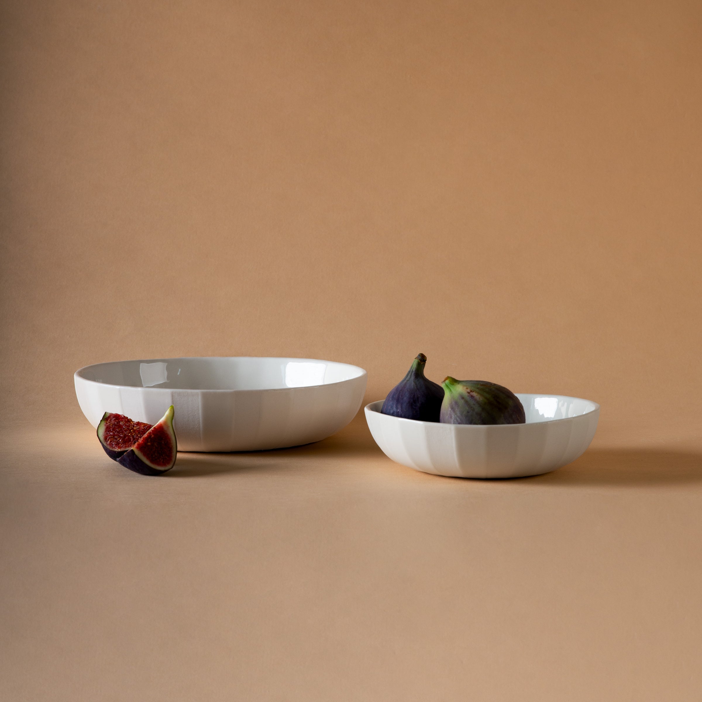 Two white ceramic bowls, part of Schneids Facet Tableware collection, rest on a neutral background. The left bowl has a cut fig beside it, and the right one is filled with whole figs. This minimalist scene exudes elegance and peace.