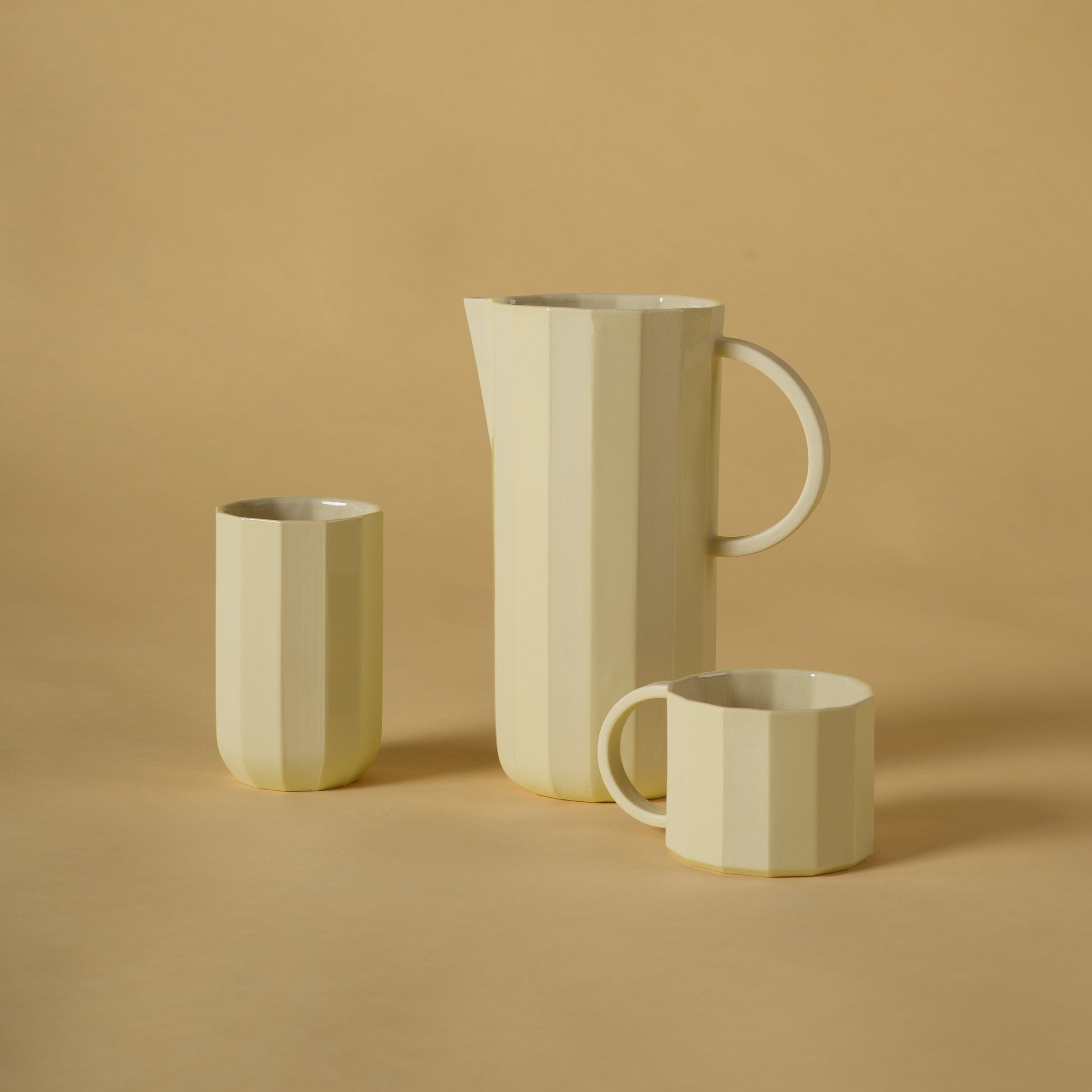 Three cream-colored ceramic pieces from Schneids Facet Tableware collection are showcased on a beige background. The set features a tall handled pitcher, a cylindrical cup, and a shorter handled mug, each highlighting intricate color dynamics in their faceted design.