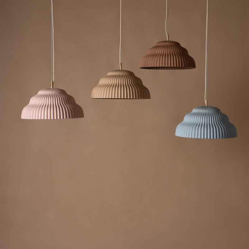 Against a beige background, four Schneids Kaskad Cloud Pink lamps with ribbed ceramic shades in pink, tan, brown, and blue hang evenly spaced. The color-matched textile cables add to the harmonious and minimalist design.