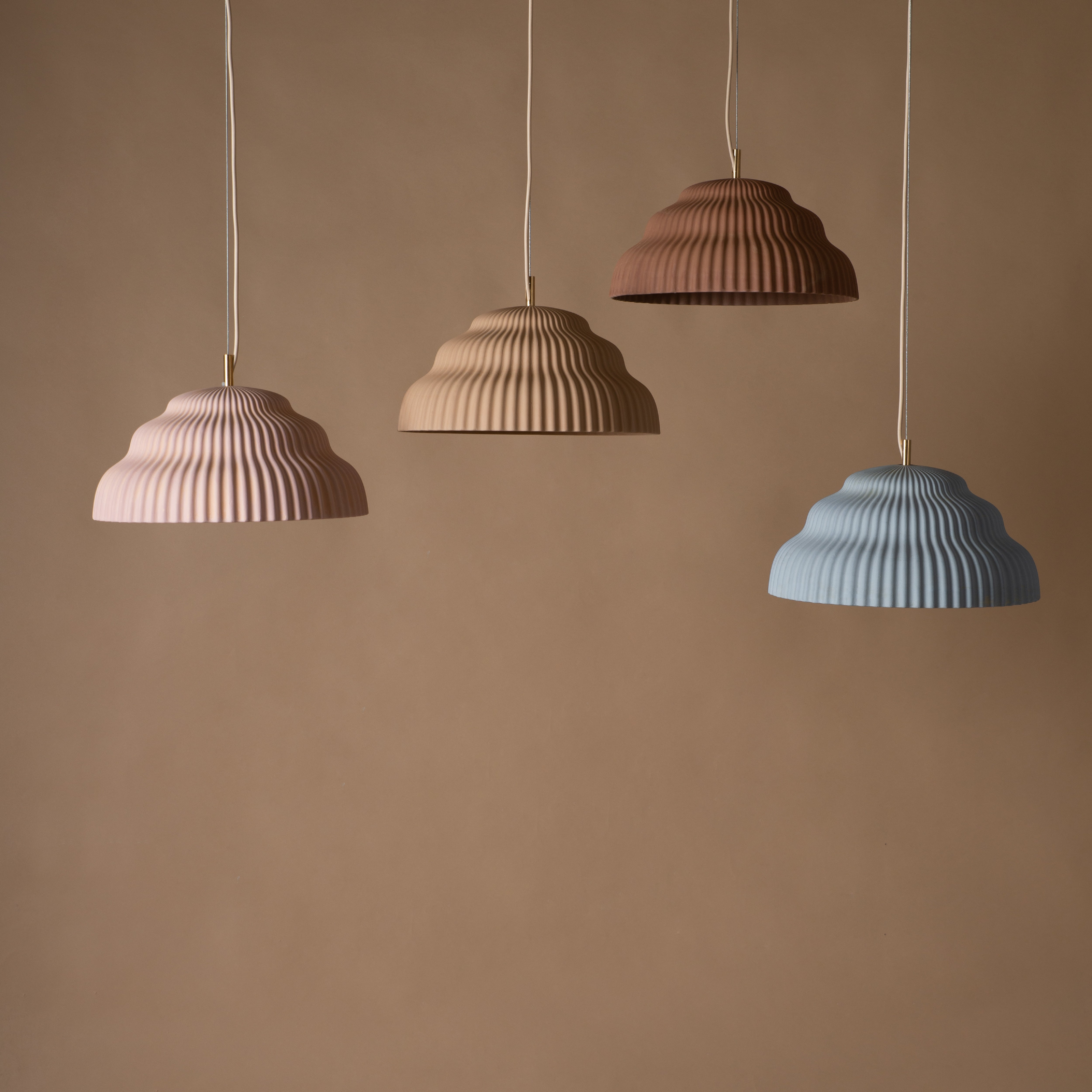 Four dome-shaped pendant lights, echoing the elegant Kaskad Biscotti design by Schneid, hang against a beige background. Their ribbed texture features shades of pink, light brown, brown, and light blue and they are suspended by thin white cords.