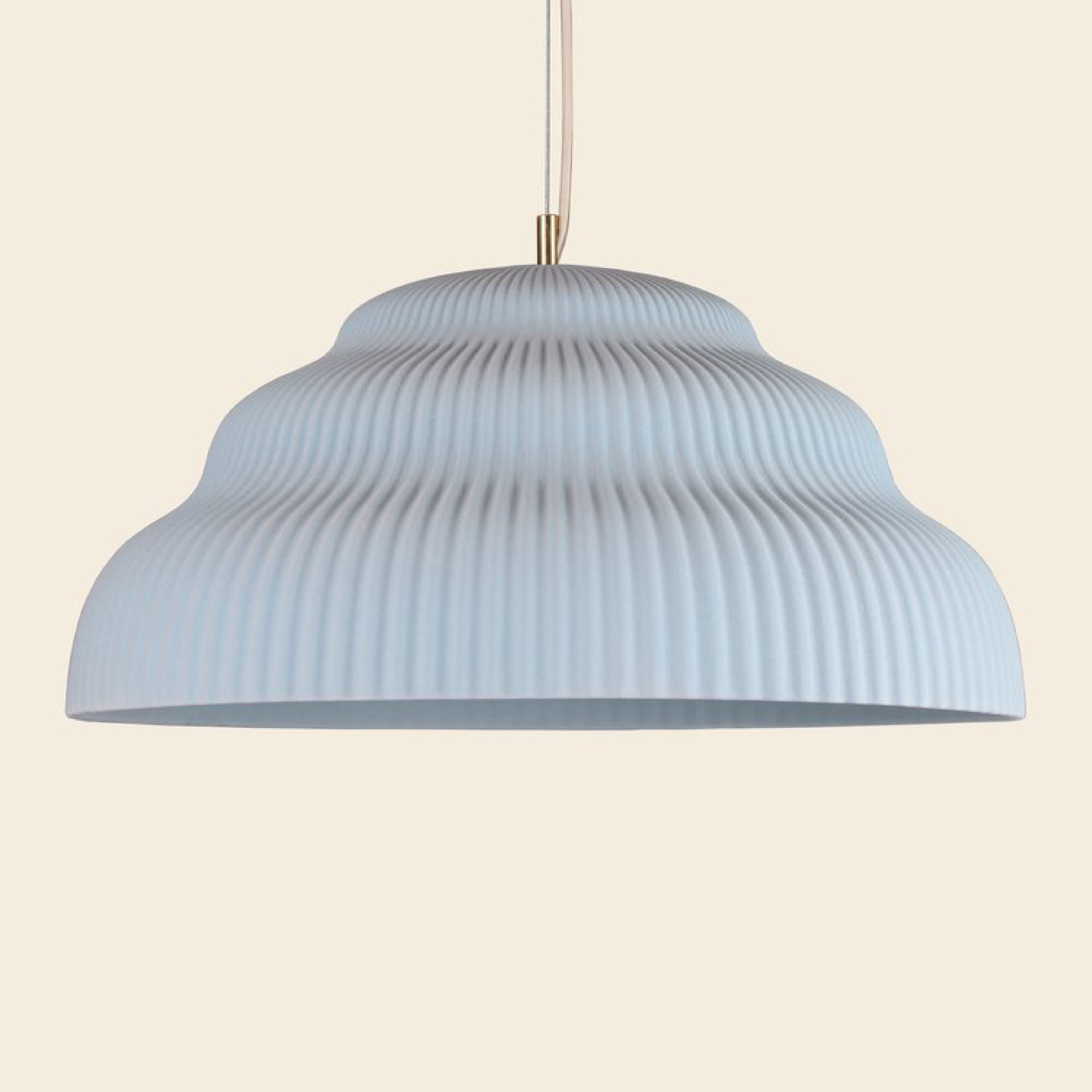 A Schneid Kaskad Light Blue pendant lamp with a ribbed, light grey shade hangs from a cord against a soft beige background. Its wavy, textured design gives this ceramic lamp an exquisite contemporary touch, making it an outstanding piece of designer lighting.