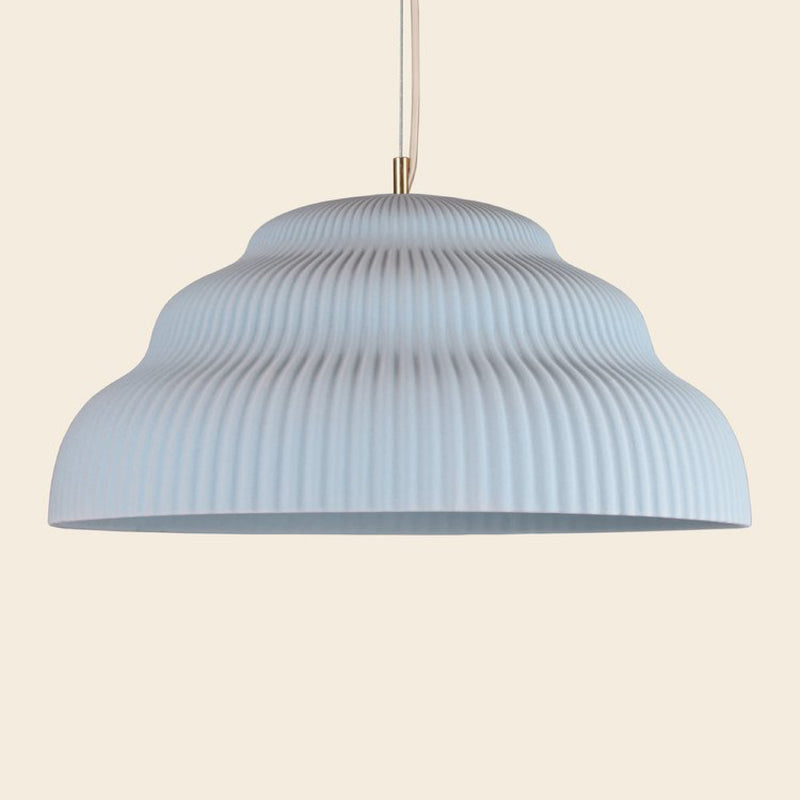 A Schneid Kaskad Light Blue pendant lamp with a ribbed, light grey shade hangs from a cord against a soft beige background. Its wavy, textured design gives this ceramic lamp an exquisite contemporary touch, making it an outstanding piece of designer lighting.