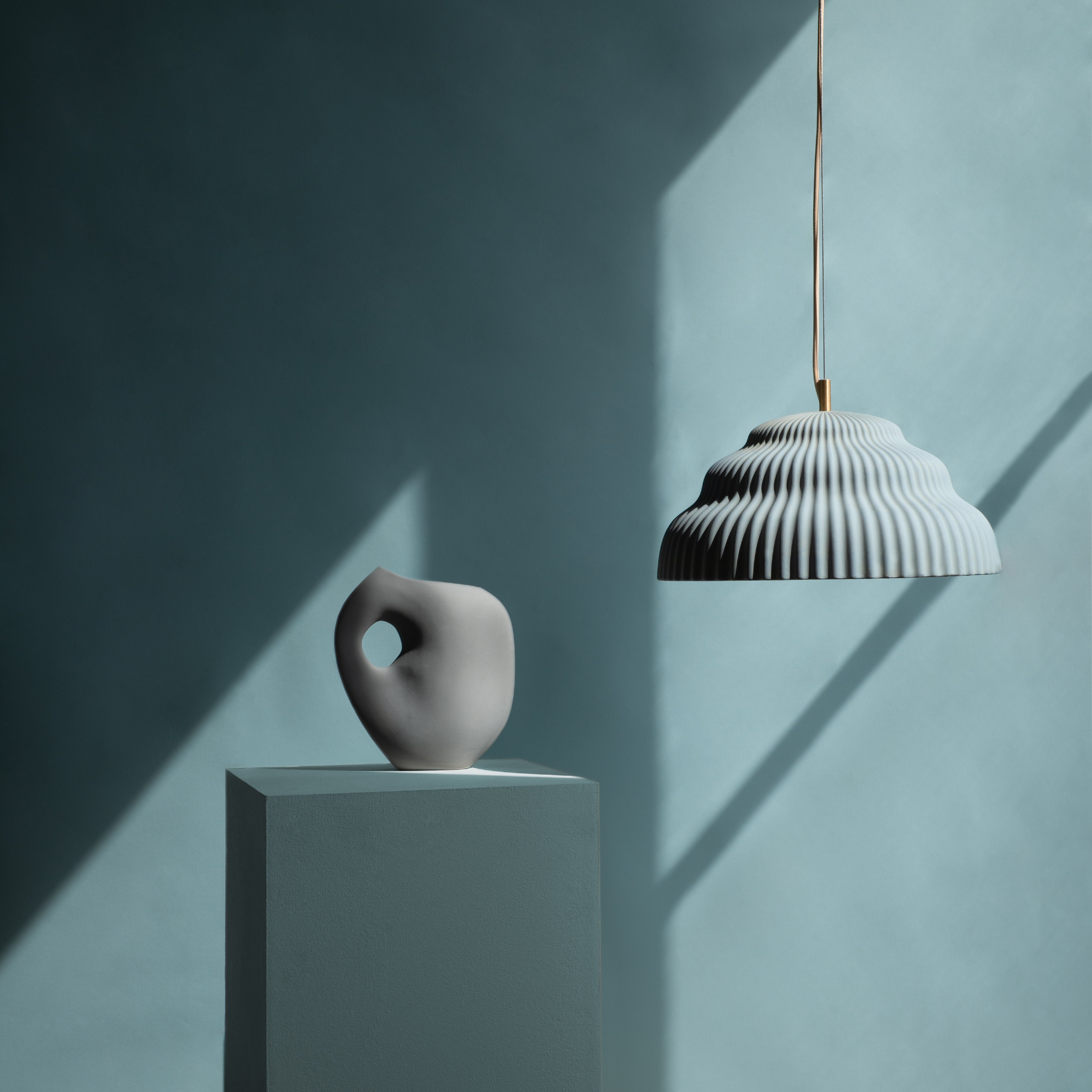 A minimalist setup features a soft light blue pedestal with a gray abstract vase, while a Schneid Kaskad Light Blue ceramic pendant lamp above casts intricate shadows on the solid background, adding designer lighting elegance.