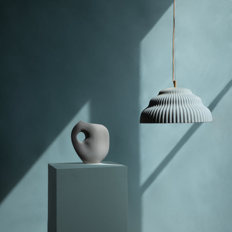 A minimalist setup features a soft light blue pedestal with a gray abstract vase, while a Schneid Kaskad Light Blue ceramic pendant lamp above casts intricate shadows on the solid background, adding designer lighting elegance.