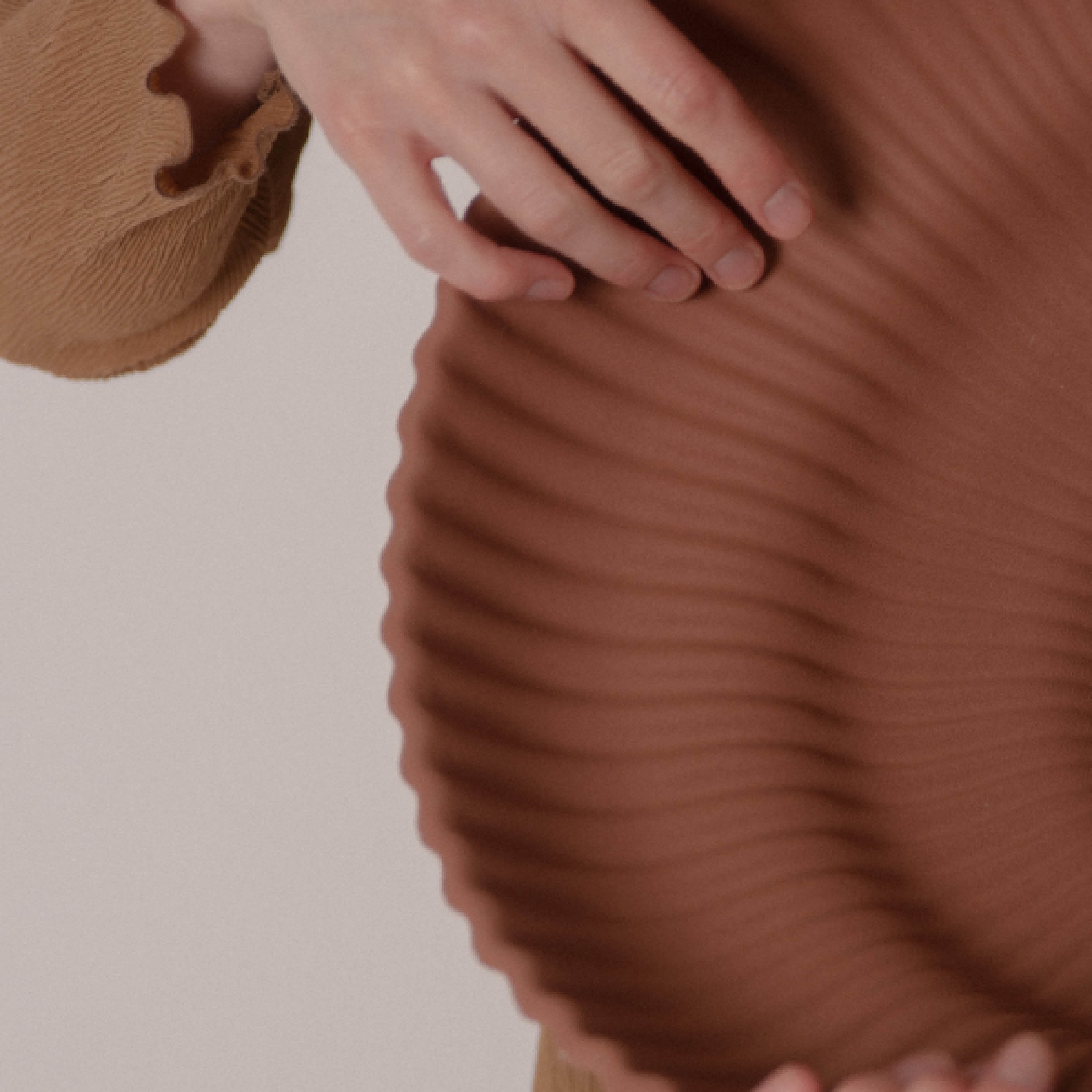 A hand touches textured, wavy, brown material like the Kaskad Biscotti lamp by Schneid. The person wears a beige long-sleeve with a ruffled cuff against a light-toned background.