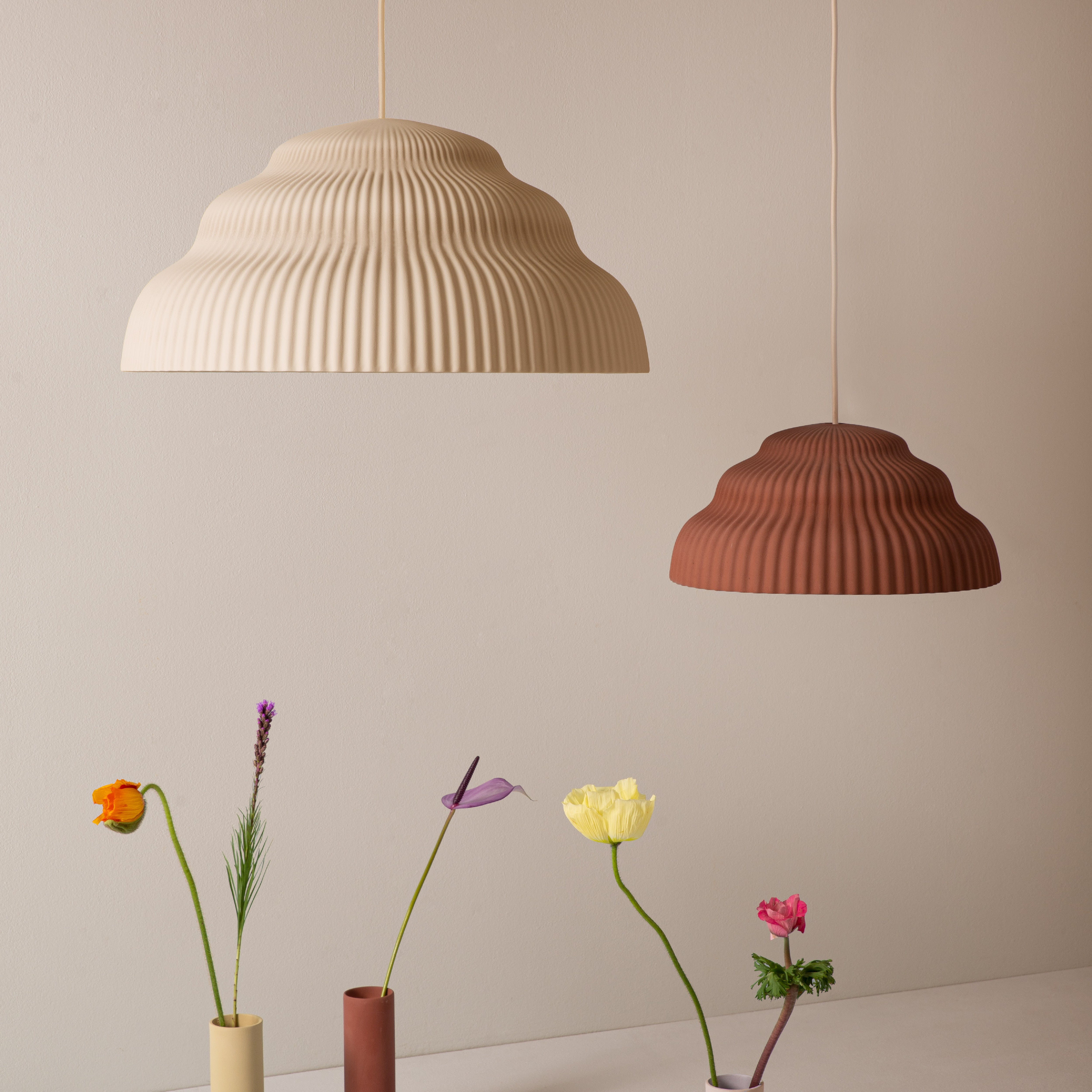 The Kaskad Biscotti ceramic suspension lamps by Schneid in beige and terracotta elegantly hover over a minimalist display of vibrant flowers in slender vases on a neutral background, highlighting their subtle beauty.
