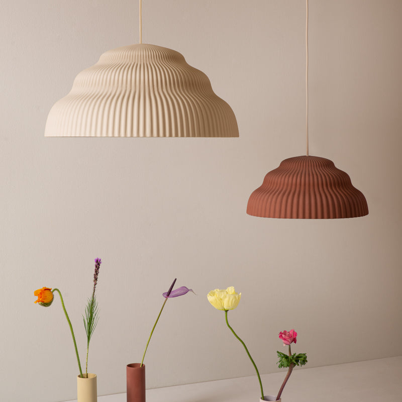 The Kaskad Biscotti ceramic suspension lamps by Schneid in beige and terracotta elegantly hover over a minimalist display of vibrant flowers in slender vases on a neutral background, highlighting their subtle beauty.