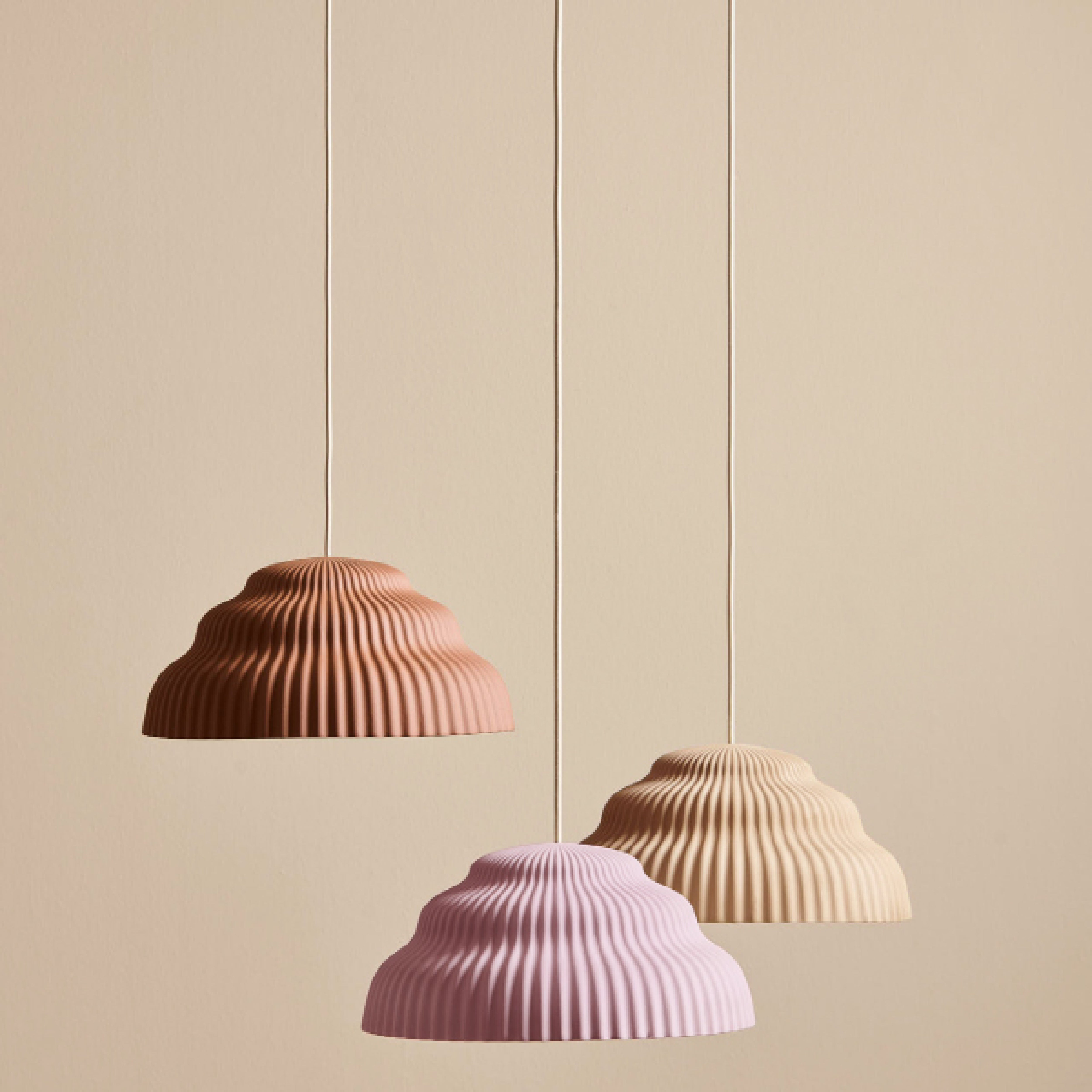 Three Schneid Kaskad Cloud Pink lamps with ribbed, scalloped shades in pink, light brown, and beige hang against a neutral backdrop. Each ceramic pendant is suspended by a matching textile cable, forming an elegant composition.
