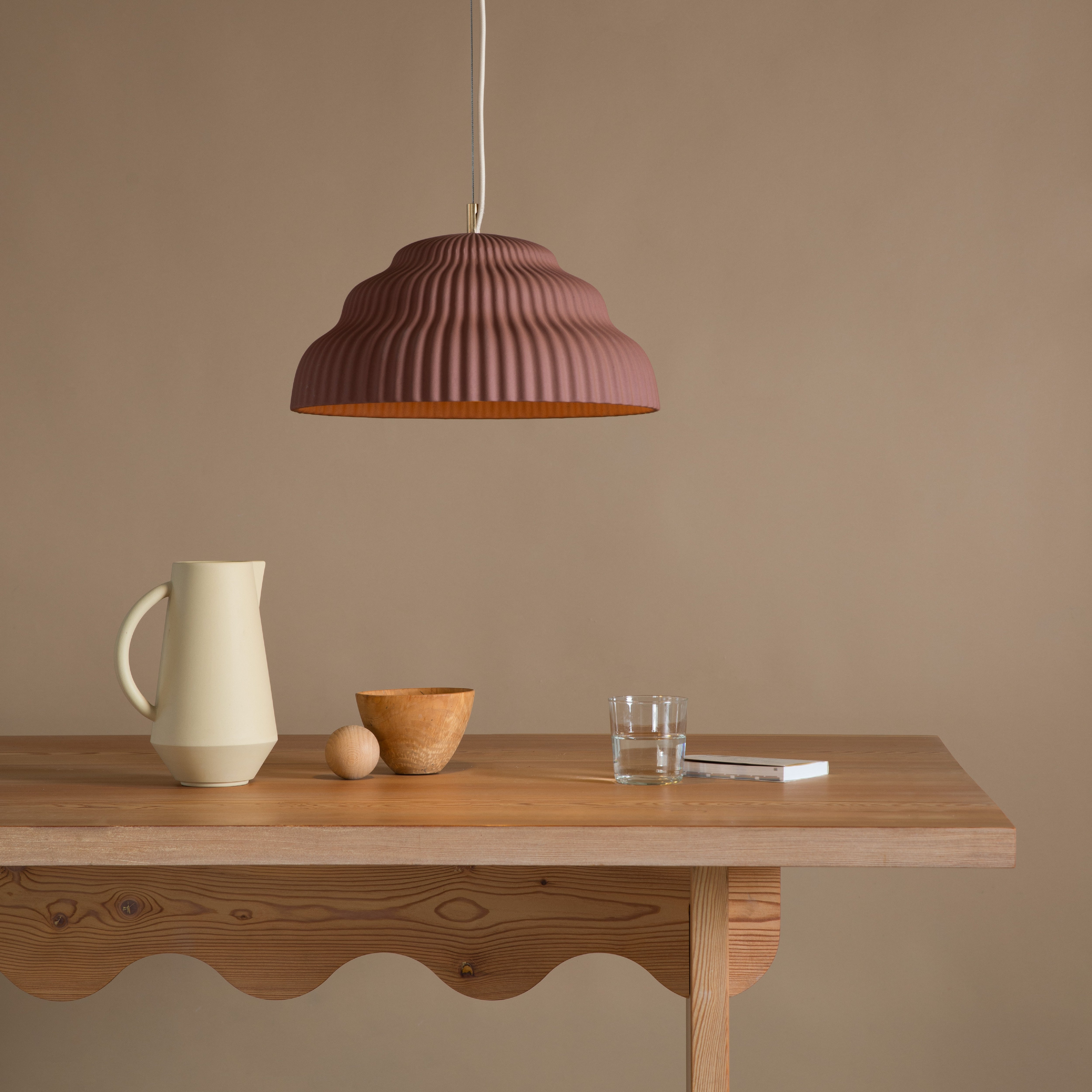 A wooden table with a scalloped edge displays a cream pitcher, small wooden bowl, round object, glass of water, and closed white book. Overhead, Schneids Kaskad Rust suspension lamp in earthy tones casts warm light against the beige background.