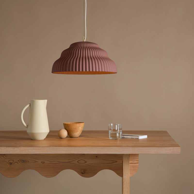 A wooden table with a scalloped edge displays a cream pitcher, small wooden bowl, round object, glass of water, and closed white book. Overhead, Schneids Kaskad Rust suspension lamp in earthy tones casts warm light against the beige background.