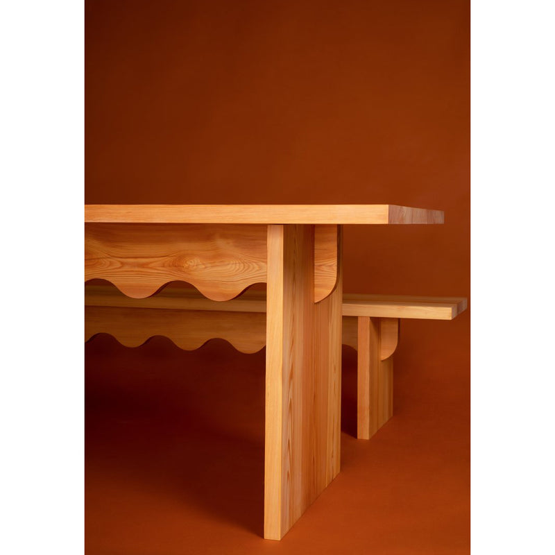 Schneids Tami Dining Collection features wooden tables with wavy edges on a warm brown backdrop. The large dining table has a scalloped underside, and the smaller bench mirrors this style with clean lines and naturally oiled larch wood grain.
