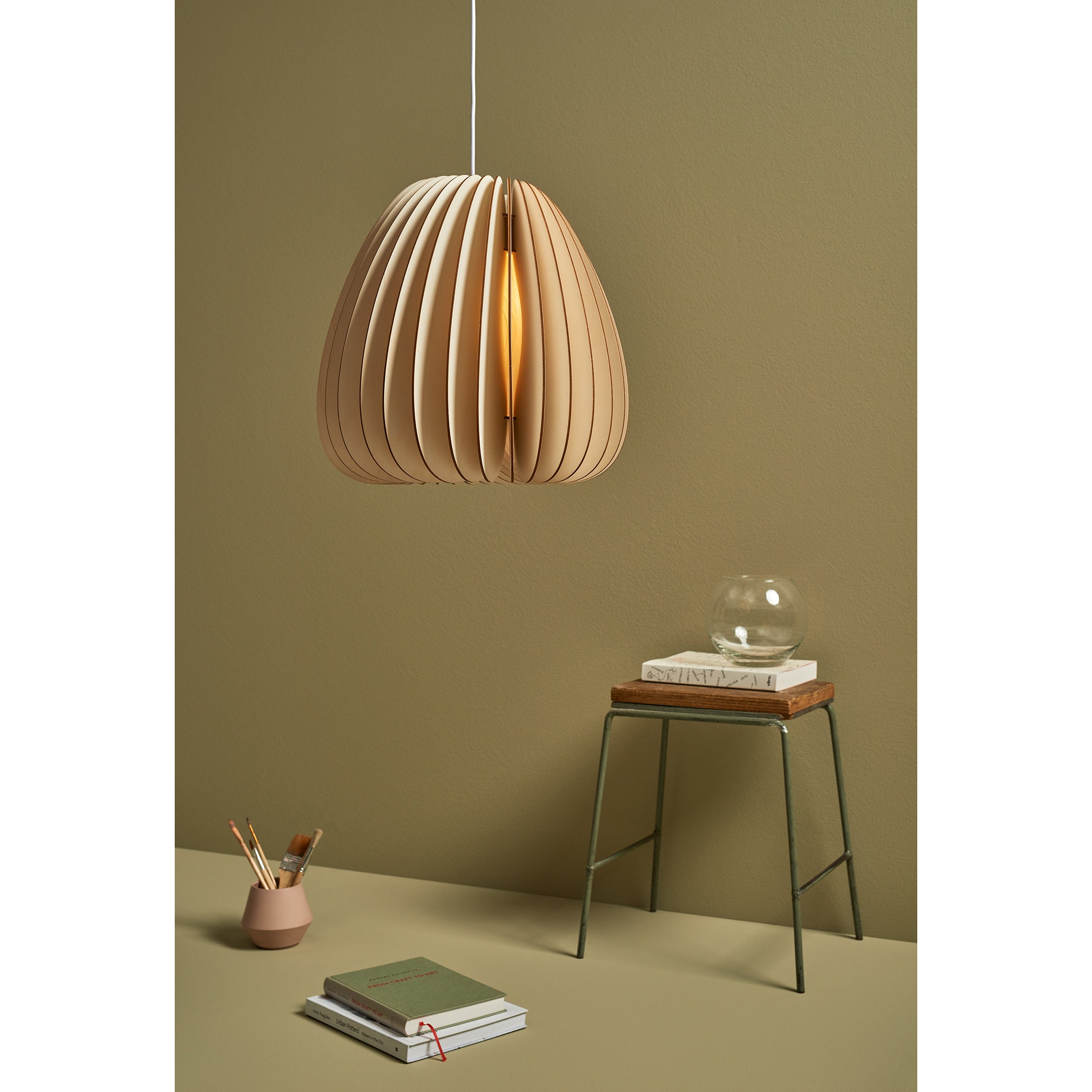 The Volum pendant lamp by Schneid, crafted with wooden slats, creates a natural ambiance hanging against an olive-green wall. Below, a small table holds books and a glass orb with a vase of brushes and pencils nearby, while a notebook rests on the floor.