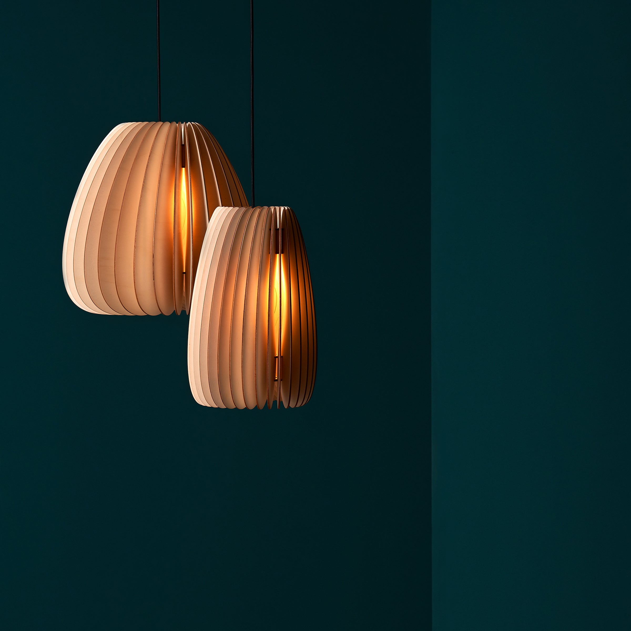 The sale items, Secundum pendant lights by Schneid, featuring modern wooden slats in an organic design, hang against a dark green wall and cast a warm glow.