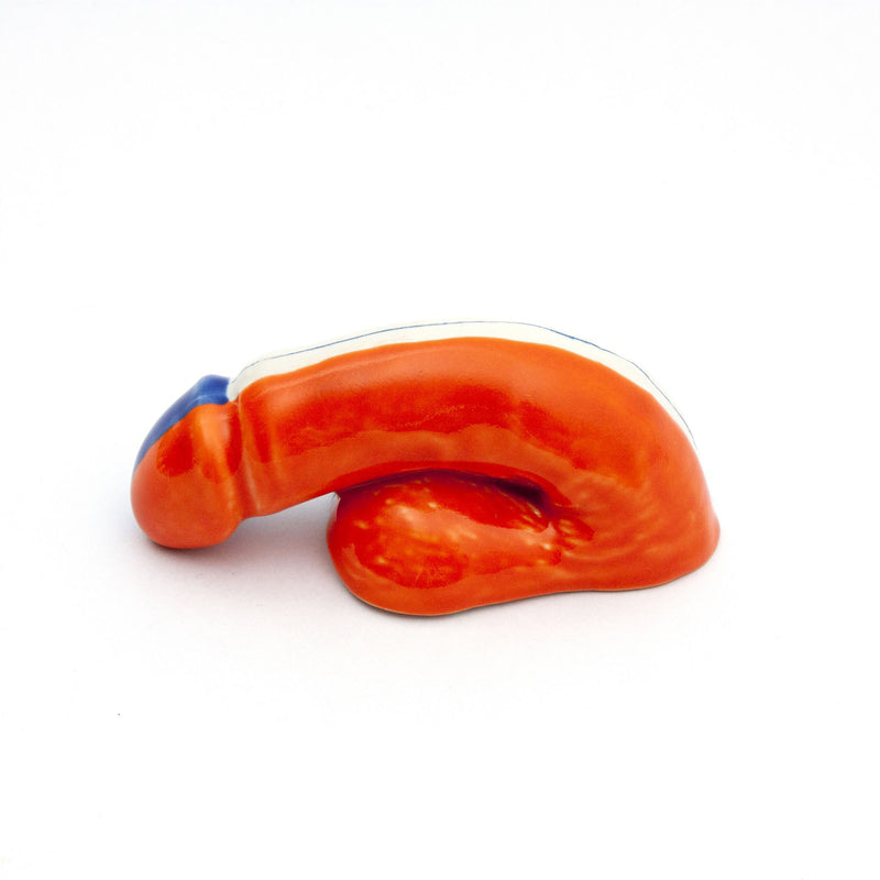 The vibrant orange A Decorative Sex - Go Mets by Schwanzmanufaktur is an Austrian handmade porcelain sculpture on a white background, boasting a smooth, elongated design with a delicate blue accent at one end.