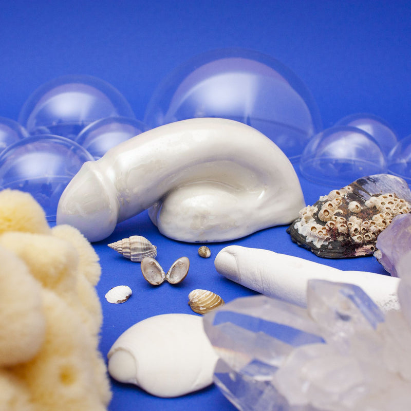 Schwanzmanufakturs A Decorative Sex - Pearl presents a captivating sea-themed display on a blue backdrop with a handmade porcelain shell, various seashells, coral pieces, barnacles, and crystals. Translucent bubbles elegantly float in the background echoing Austrian craftsmanship.