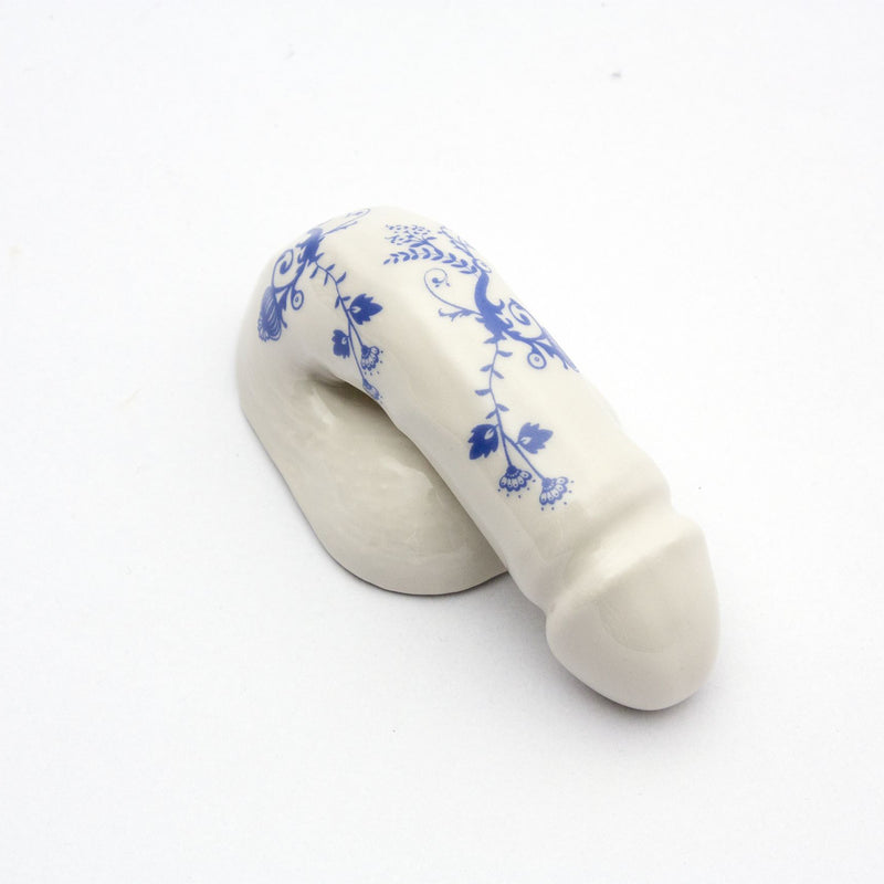 The Decorative Sex - Blue Onion 3 by Schwanzmanufaktur is a porcelain sculpture with a phallic form, featuring white and blue floral designs, elegantly resting on a surface—an exquisite piece echoing Oscar Wildes wit in its bold yet refined presentation.