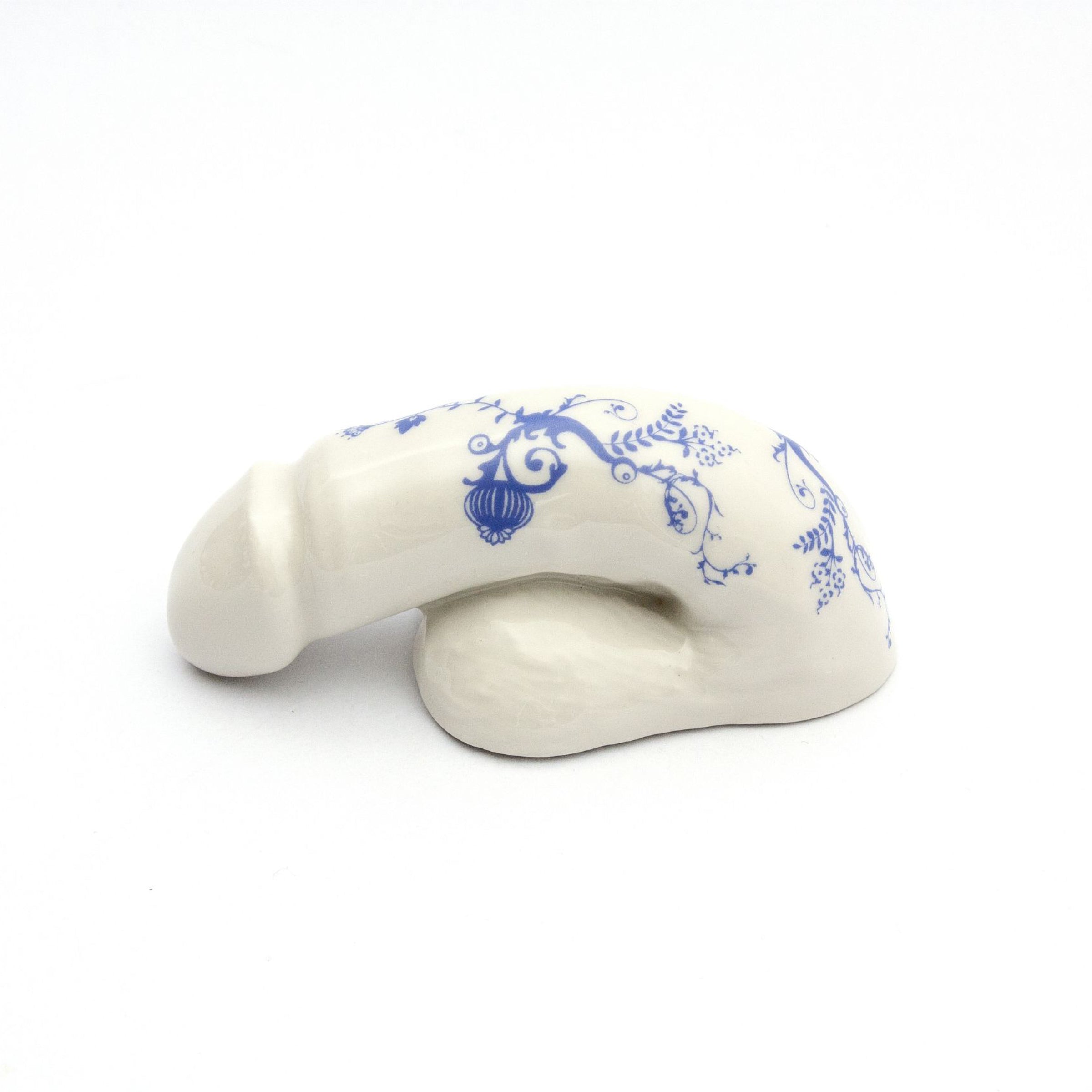 The A Decorative Sex - Blue Onion 3 by Schwanzmanufaktur is a white ceramic sculpture shaped like a banana with intricate blue floral designs, reminiscent of porcelain, and elegantly rests on a smooth white surface.